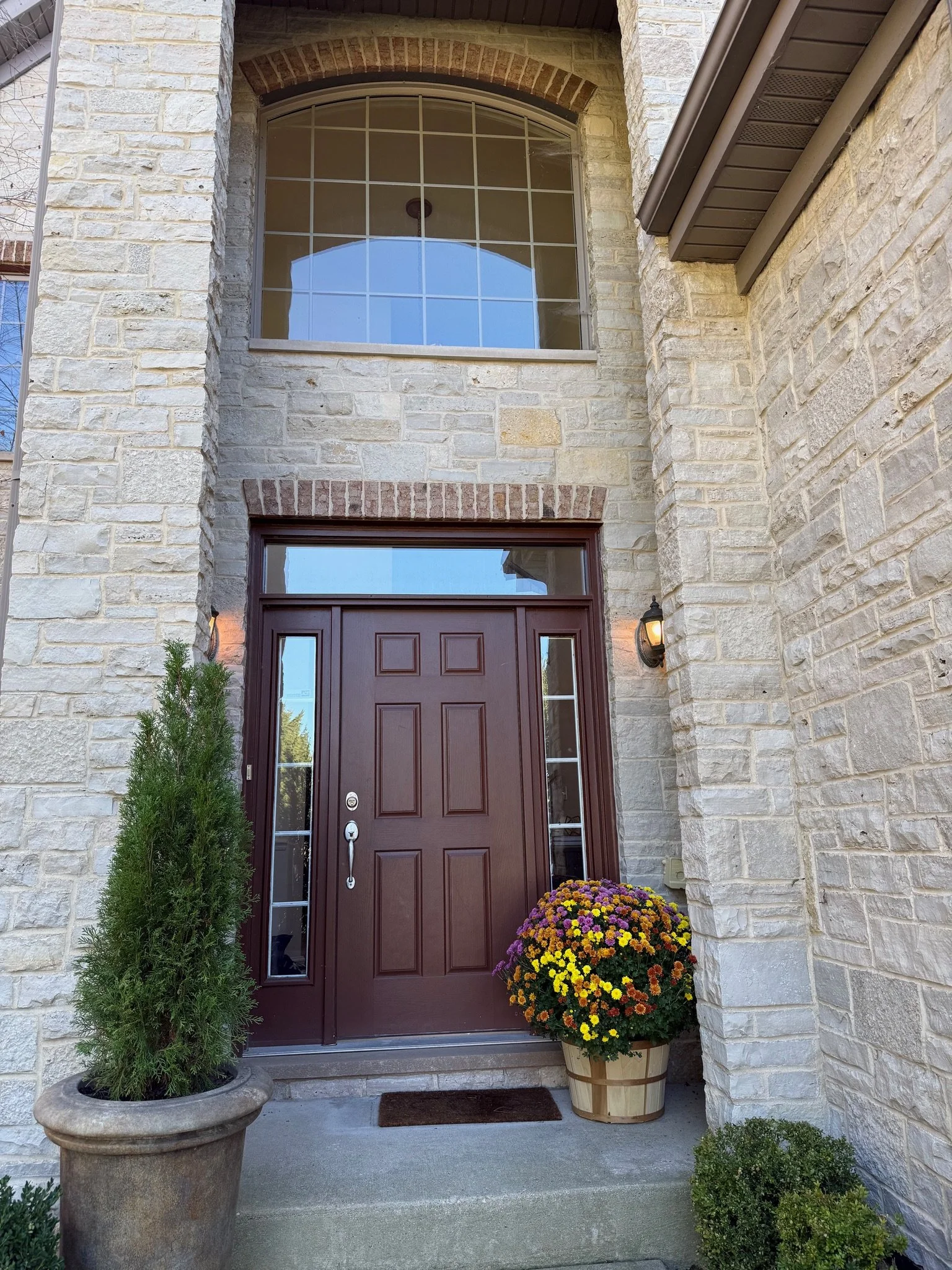 Front door of a house with a brick and stone exterior, a large window above the door, and potted plants including a tall green bush and a colorful flower arrangement on either side of the door.