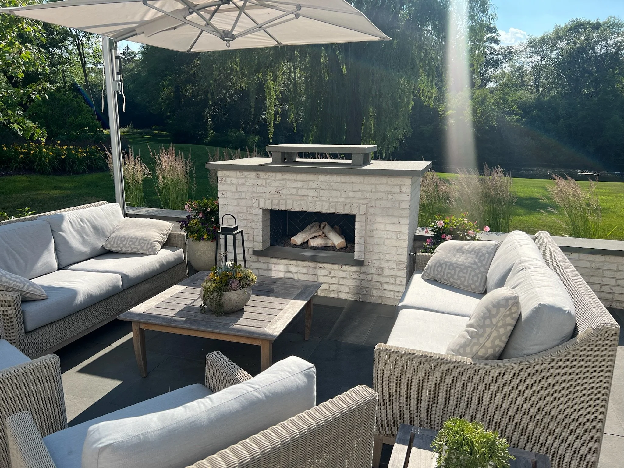 Outdoor patio area with wicker furniture featuring white cushions, a white brick fireplace, a wooden coffee table with a pot of succulents, and a large umbrella, all set in a lush green garden with trees and a pond in the background.