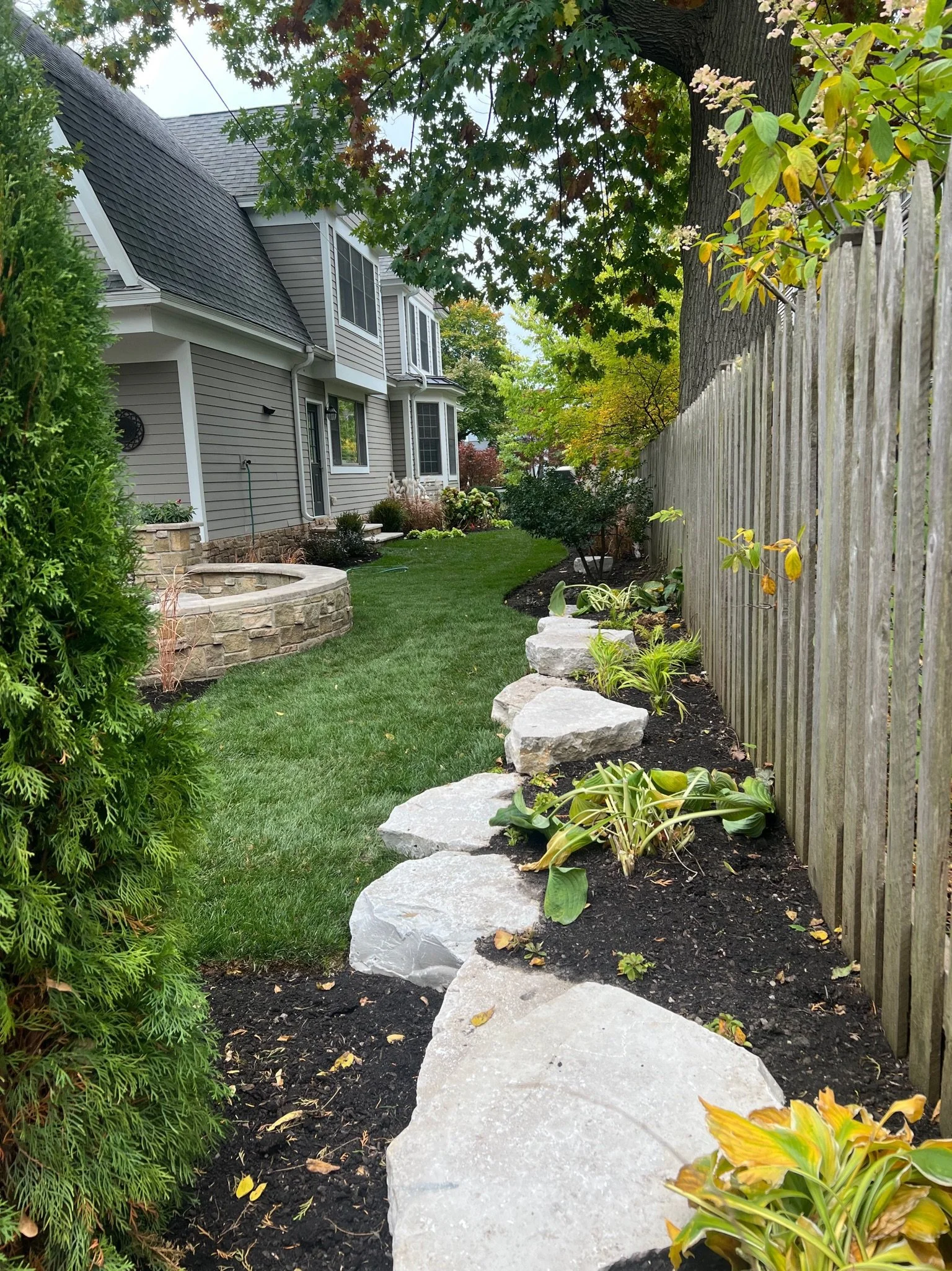 A backyard garden with a house on the left, a wooden fence on the right, green grass, plants, trees, and a stone pathway.