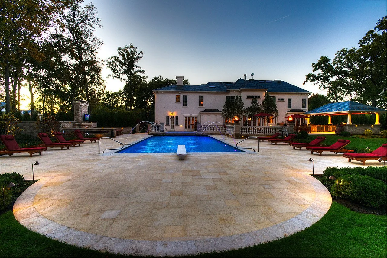 Backyard with a swimming pool, surrounded by lounge chairs and trees, at dusk.