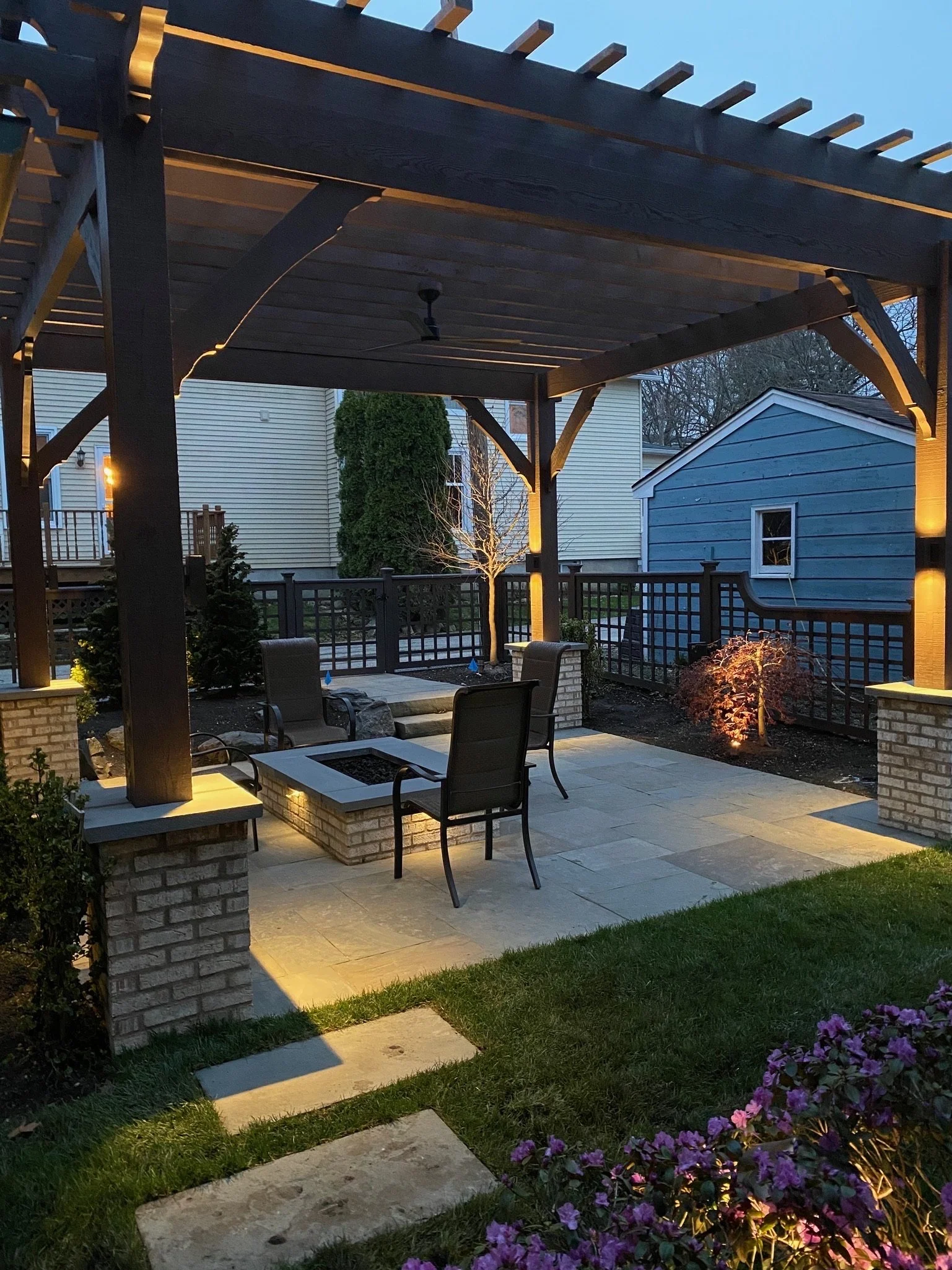 An outdoor patio area illuminated by lights, featuring a wooden pergola, patio furniture, a small fire pit, and a garden with purple flowers and trees.