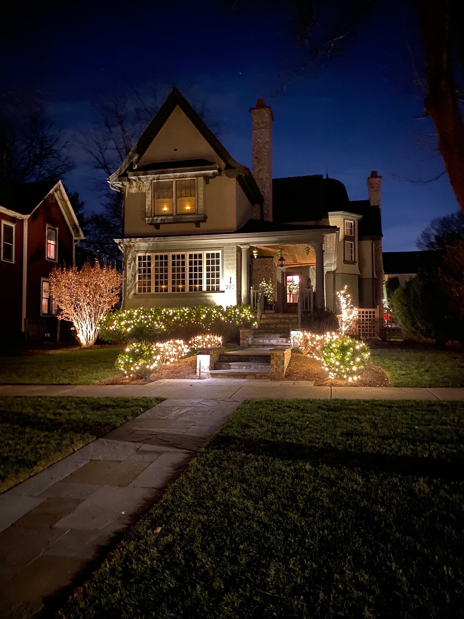 A two-story house decorated with Christmas lights and ornaments, illuminated at night with a clear dark blue sky and stars overhead. There are trees and a well-kept lawn in front of the house.