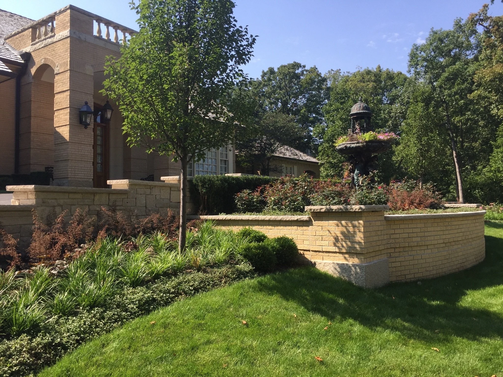 A landscaped garden with green grass, shrubs, and small trees in front of a brick house. There is a fountain with a statue, surrounded by plants, and a stone wall border.
