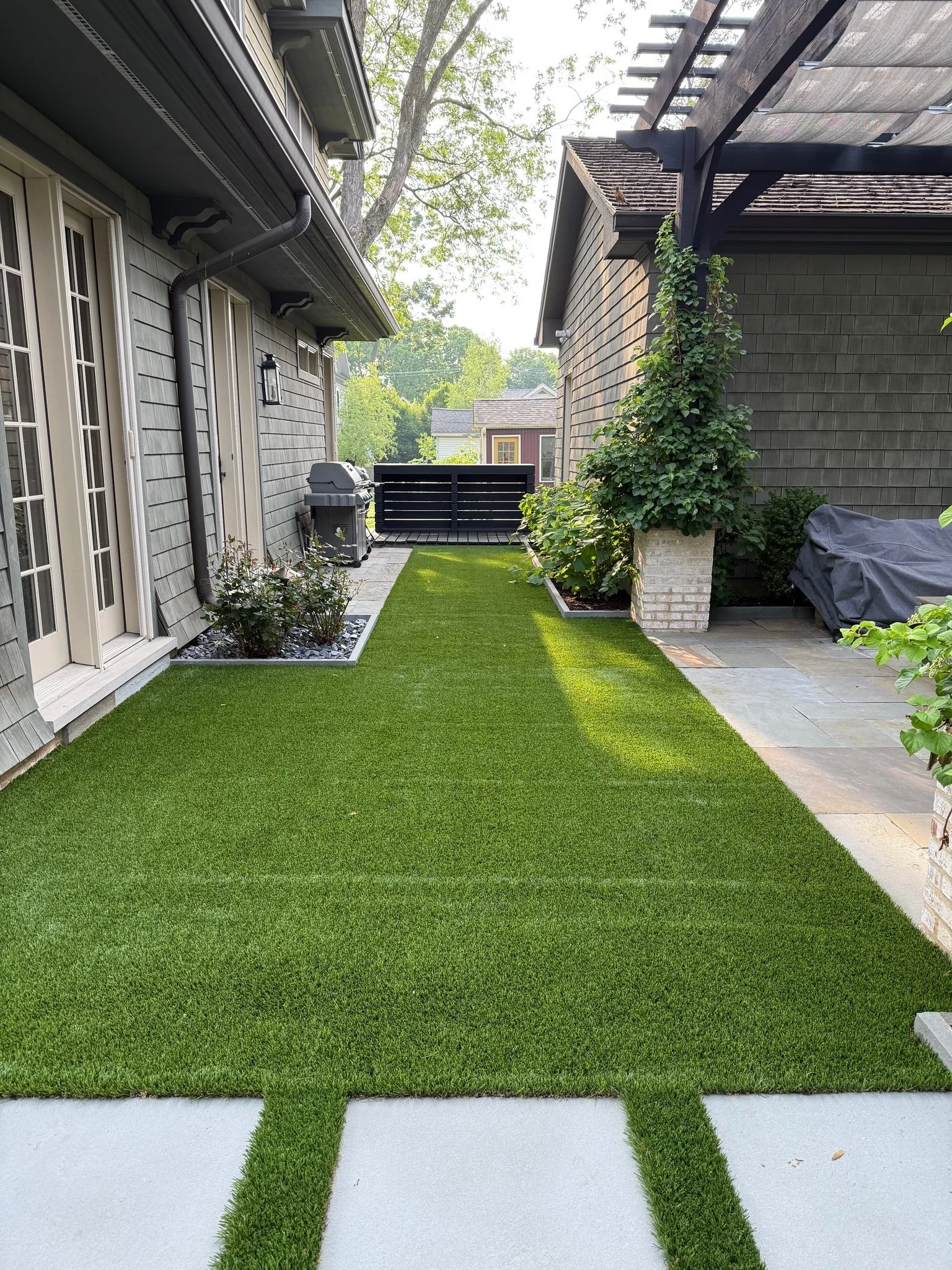 Backyard with artificial grass and white concrete pathway, house with beige siding, black shutters, and a gray door, plants along the edges, barbecue grill, and a wooden deck with black railing.