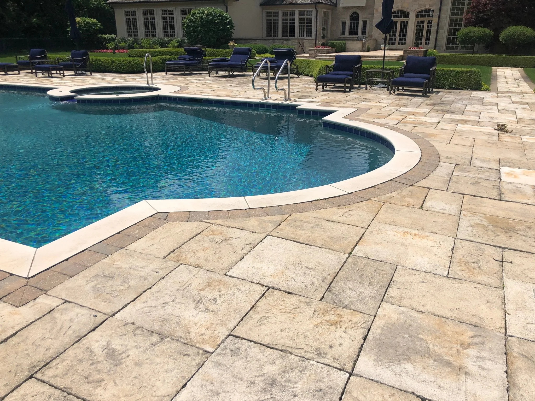 A backyard scene featuring a swimming pool with steps and a hot tub, surrounded by a stone patio with outdoor chairs and tables, green hedges, and a large house in the background.