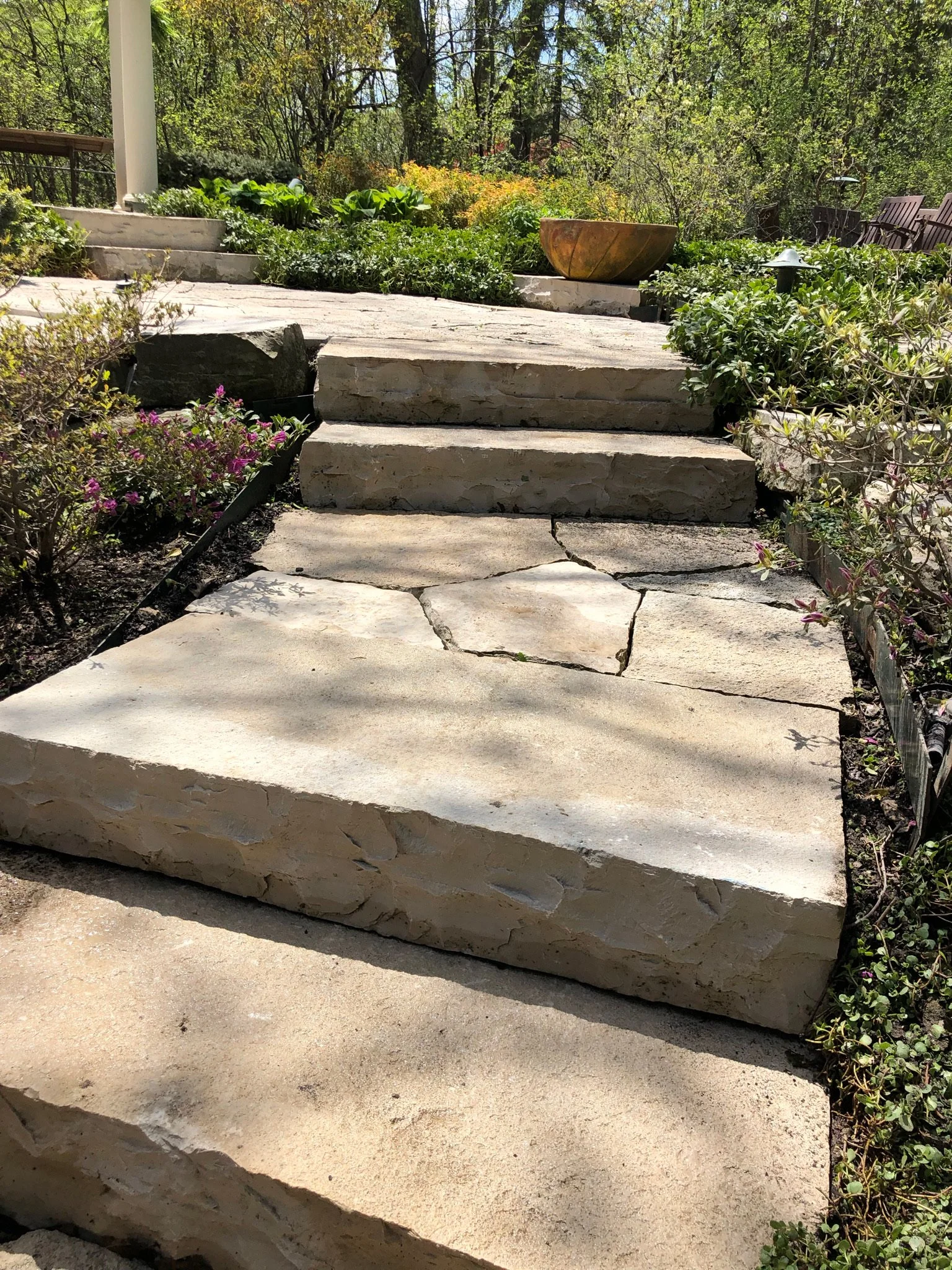Stone steps and a pathway leading upward through a garden with greenery, flowering plants, and a large decorative bowl on the top step, surrounded by trees and outdoor benches.