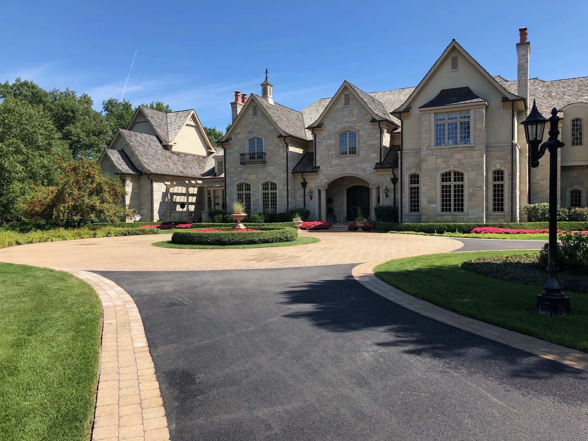 A large, elegant house constructed with stone and siding, featuring multiple gabled roofs, tall windows, and a well-maintained front yard with a driveway, garden, and decorative lamp post.