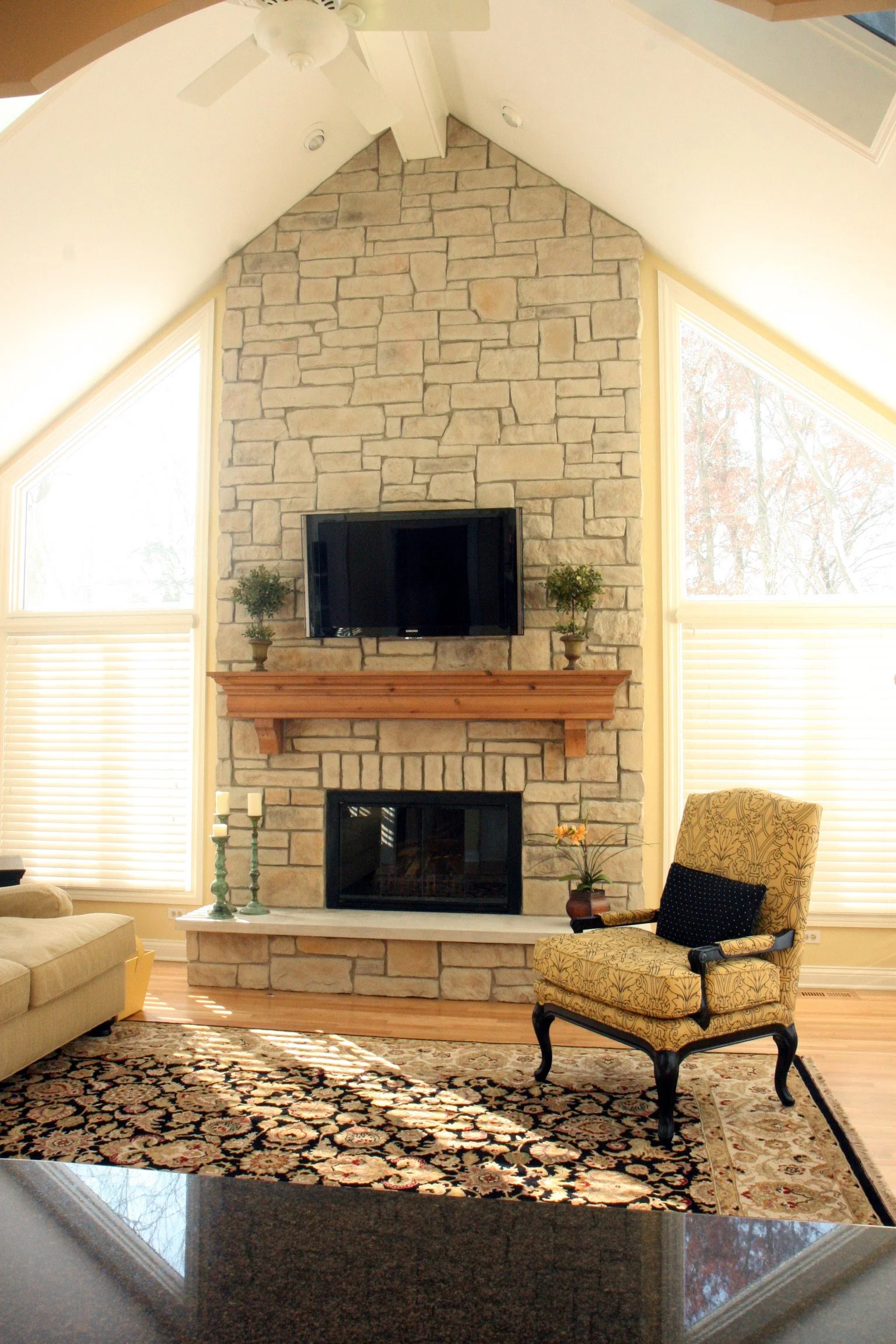 Living room with stone fireplace, TV above mantle, armchair with decorative pillow, patterned area rug, and large windows with blinds.