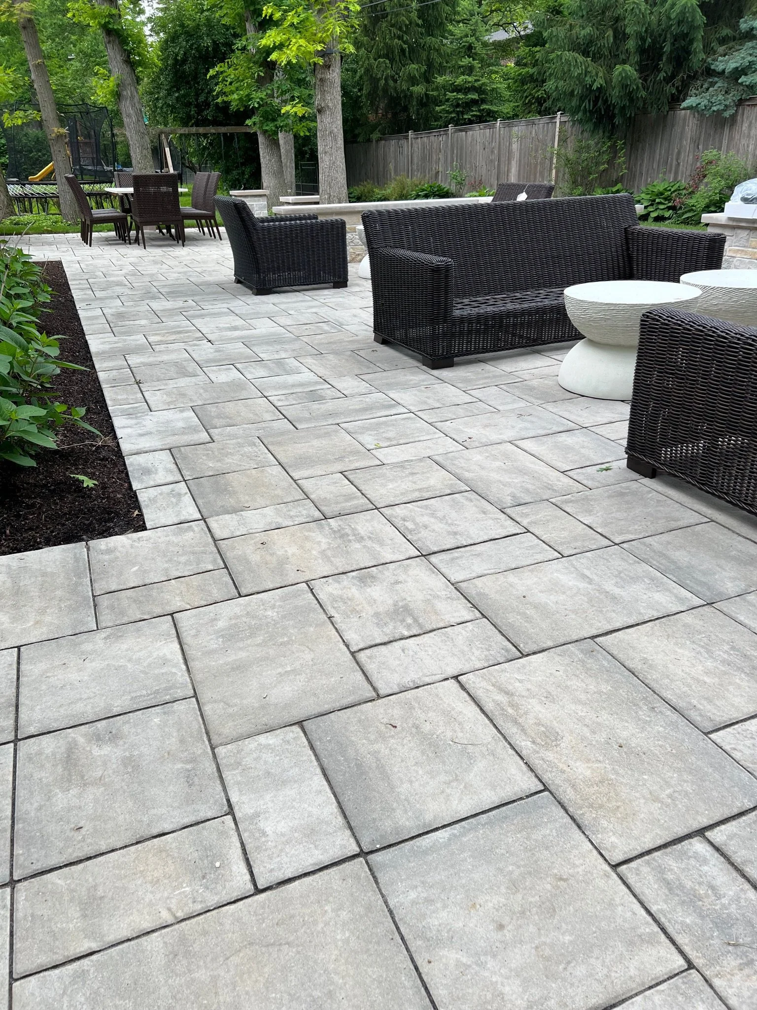 A backyard patio with light gray stone pavers, black wicker outdoor furniture including a sofa and chairs, white round side tables, surrounded by green trees and plants, with a wooden fence in the background.