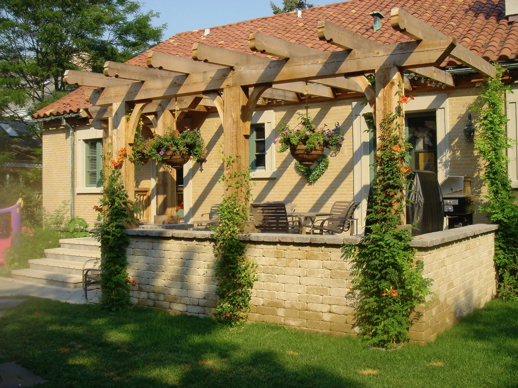 A brick house with a red tile roof and a wood pergola with hanging flower baskets. There are steps leading up to the patio area with outdoor furniture and a grill. Green plants and trees surround the house.