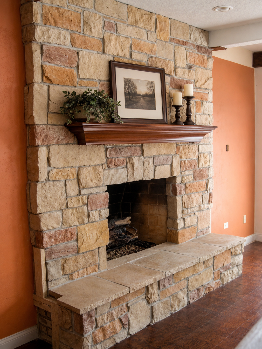 Living room fireplace with multicolored stone surround and hearth, topped with a wooden mantel decorated with a framed picture, two candle holders with candles, and a plant, against orange and white walls.