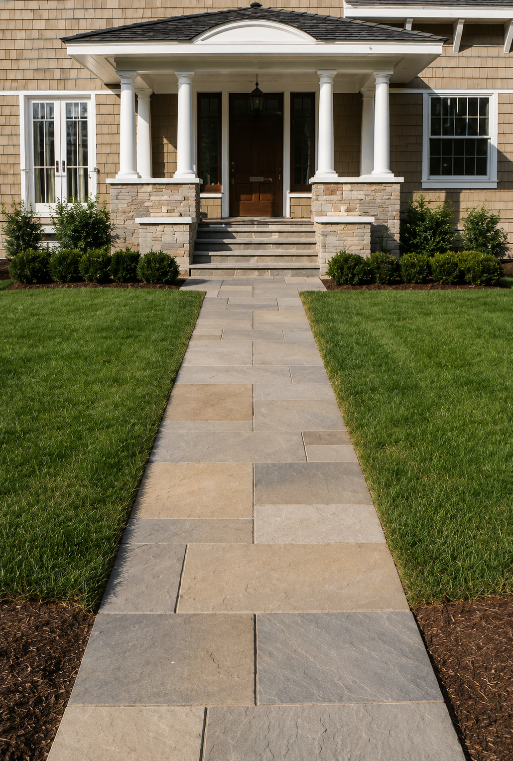 Front view of a house with a wide walkway leading to the front porch, which has four white columns, a dark wooden door, and stone steps. The house has beige siding and white-framed windows, with green grass and neatly trimmed bushes on each side of t