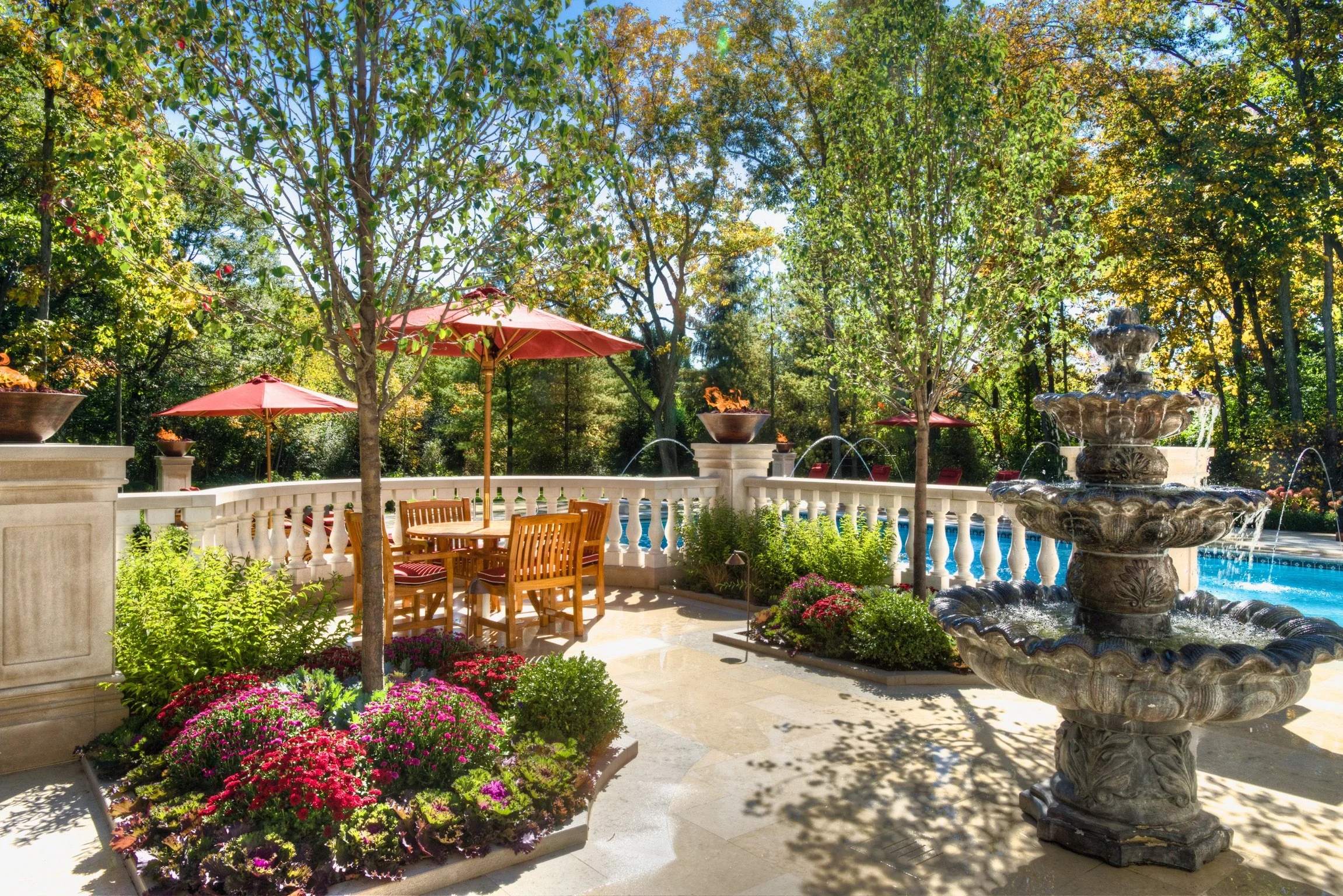 An outdoor patio area with a stone fountain, pink umbrellas, a wooden table and chairs, lush trees, colorful flowers, and sunny weather.