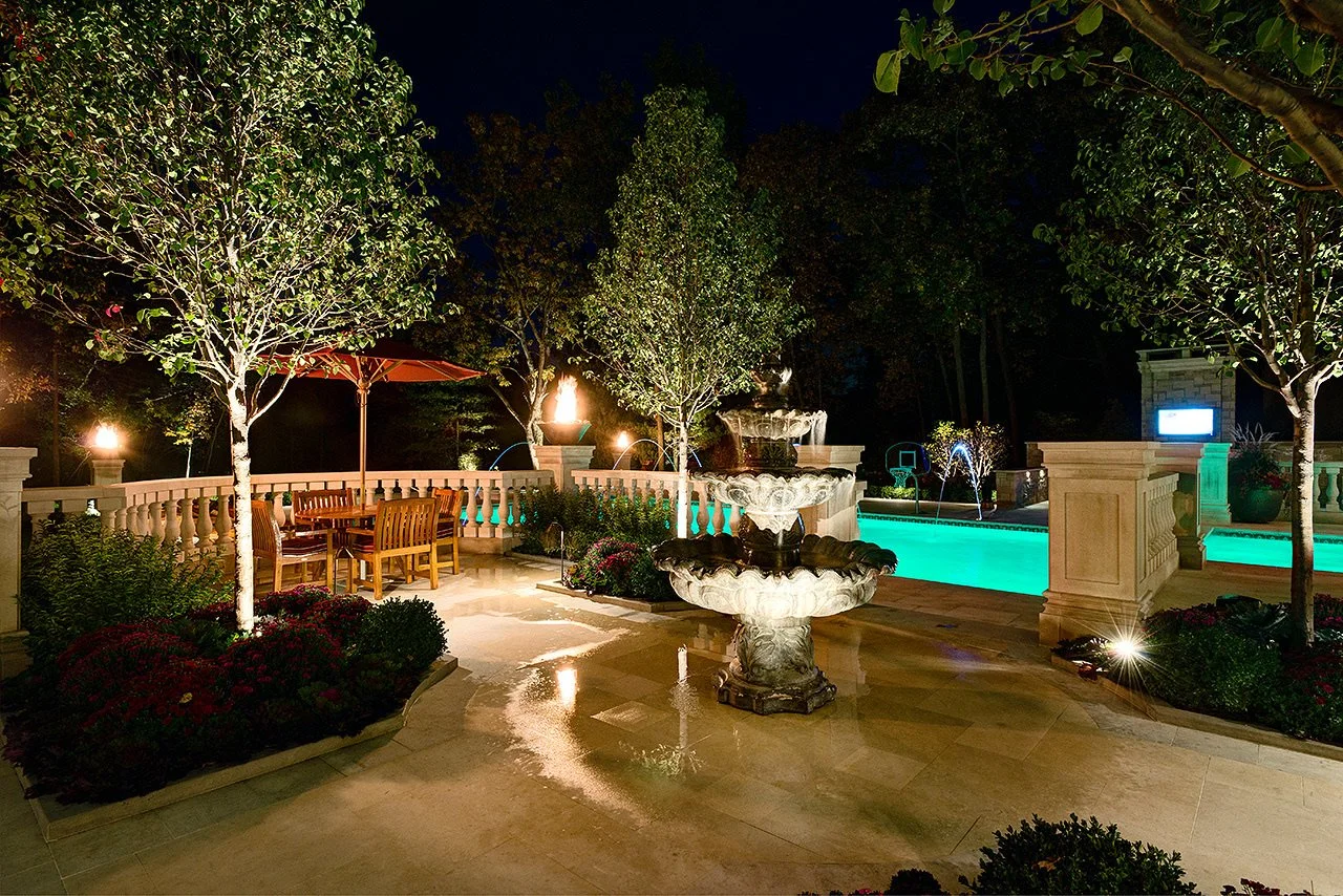 Night view of a backyard with a stone fountain, a covered dining area with a wooden table and chairs, a swimming pool with lounge chairs, trees, and landscape lighting.