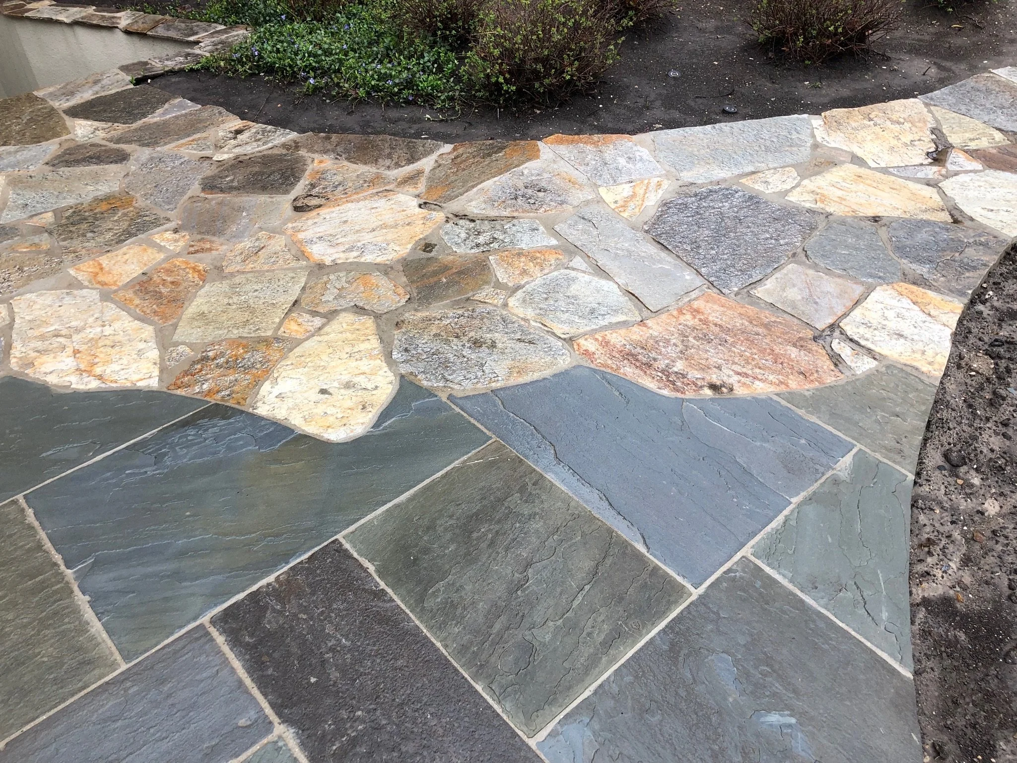A stone patio with irregularly shaped, multicolored stones in front of a garden bed with plants and soil.