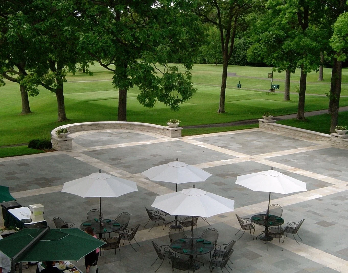 Outdoor patio with tables and white umbrellas, overlooking a grassy park with trees and walking paths.