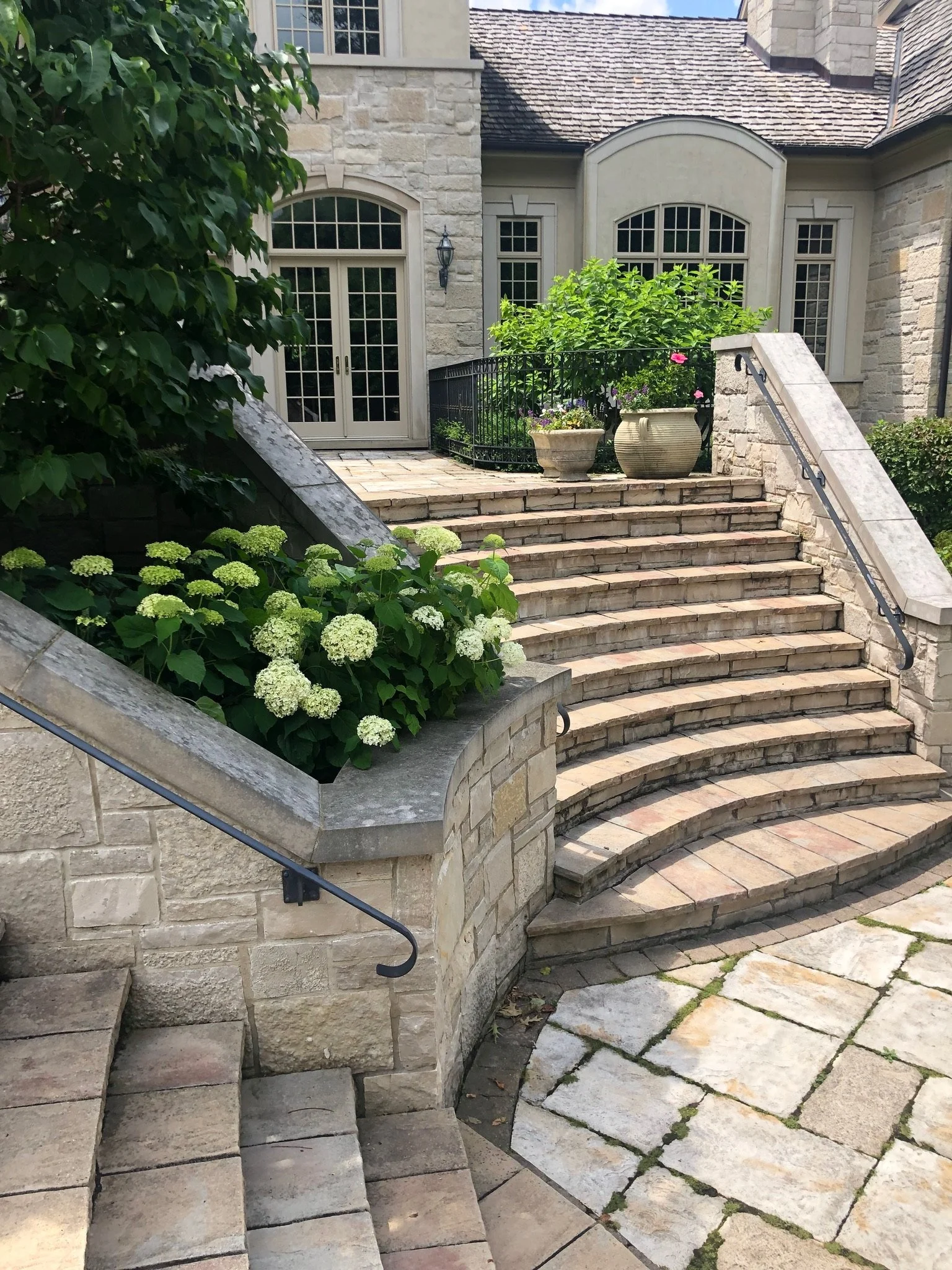 Stone stairs with black metal handrails leading up to a small porch area in front of a house with large windows and a stone exterior, surrounded by greenery and potted plants.