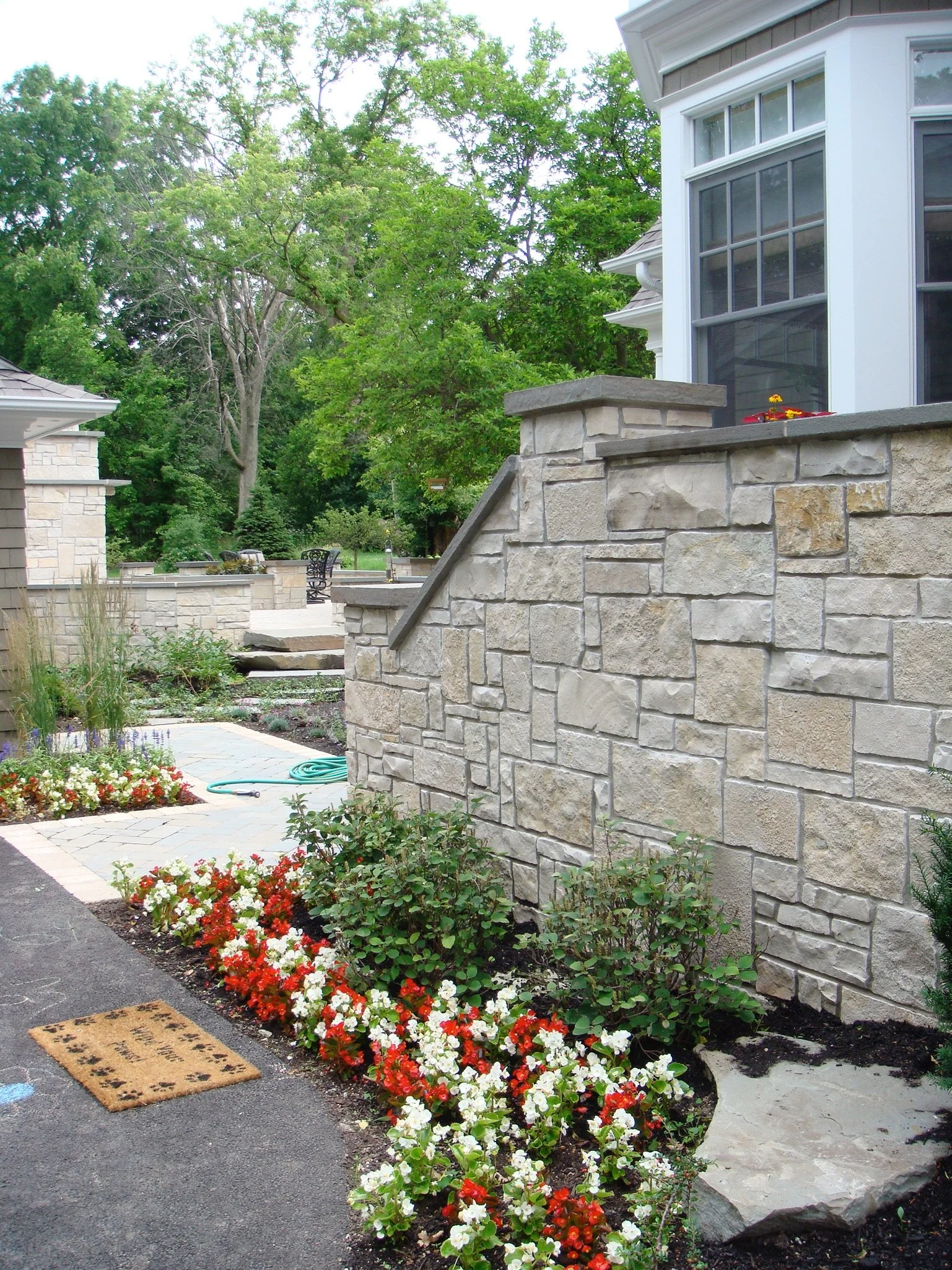A landscaped backyard patio with a stone retaining wall, colorful flower beds, a garden hose, and outdoor furniture in a lush green setting.