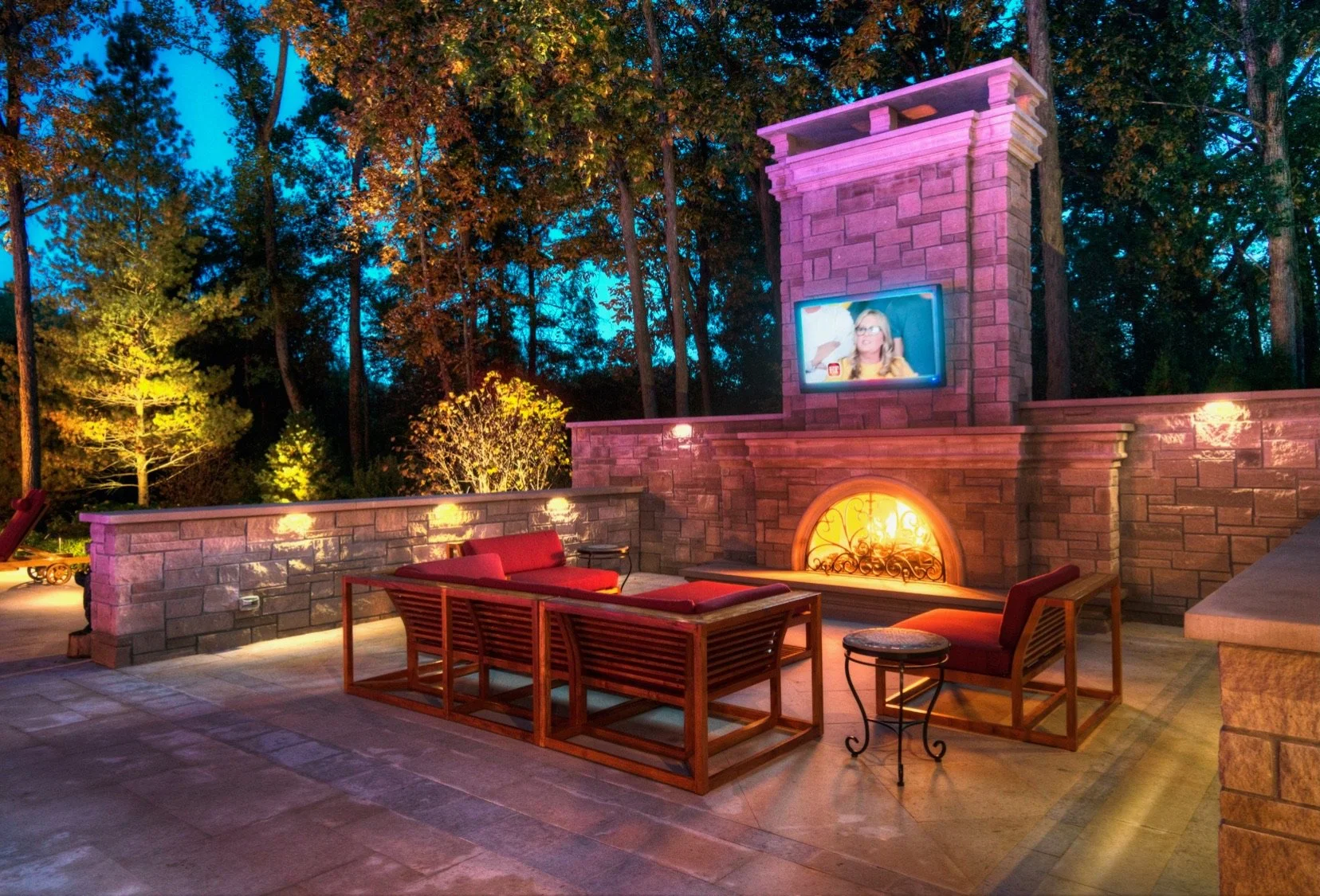 Outdoor patio area at dusk with red cushioned seating, a stone fireplace with a lit fire, a mounted television showing a woman with glasses, and tall trees in the background illuminated by outdoor lighting.