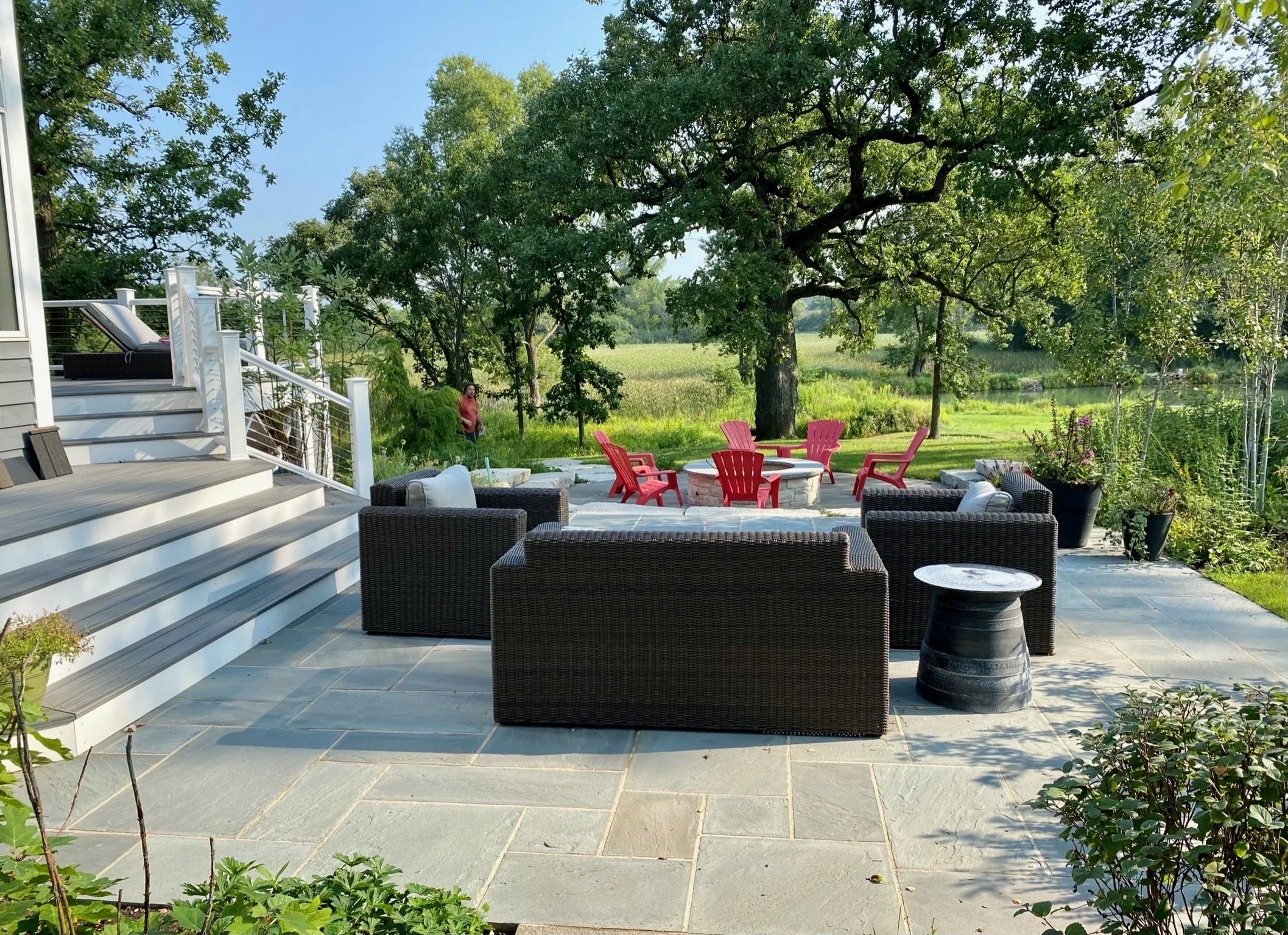 A backyard patio with outdoor furniture including a dark wicker sofa, red Adirondack chairs around a fire pit, and a black barrel-style table. A set of stairs leads up to a deck with a lounge chair, and lush green trees and open grassy fields surroun
