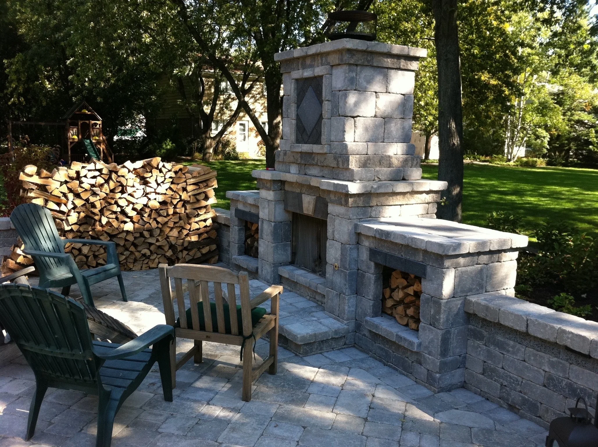 Backyard patio with a stone fireplace, stacked firewood, Adirondack chairs, and a children's playset in a green, leafy yard.