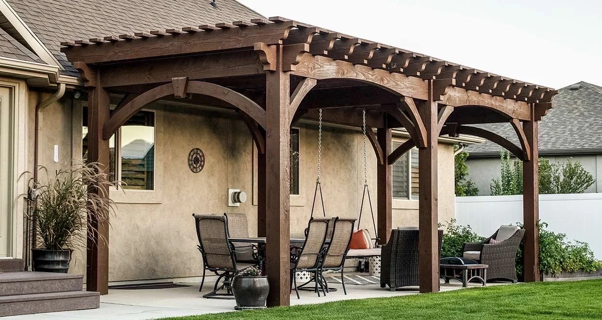 Backyard patio with a wooden pergola, outdoor dining table, chairs, a swing, and lounge chairs with cushions, surrounded by a green lawn and neighboring houses.