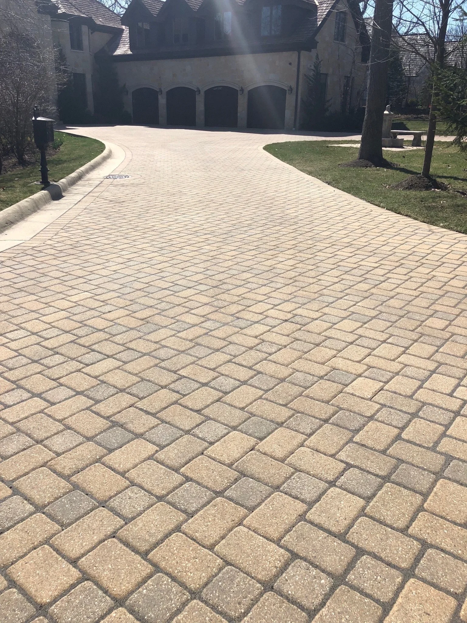 Paved driveway leading to a stone house with four garage doors, trees on the lawn, and a small bench with a water fountain nearby.