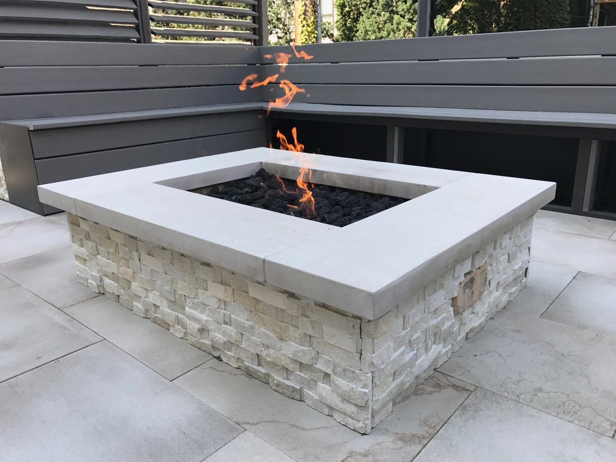 A square outdoor fire pit with flames, constructed with a white concrete border and a stone base, situated on a paved patio area with gray tiles. Surrounding it are gray wooden benches and a wooden backrest with slats, with greenery visible in the ba