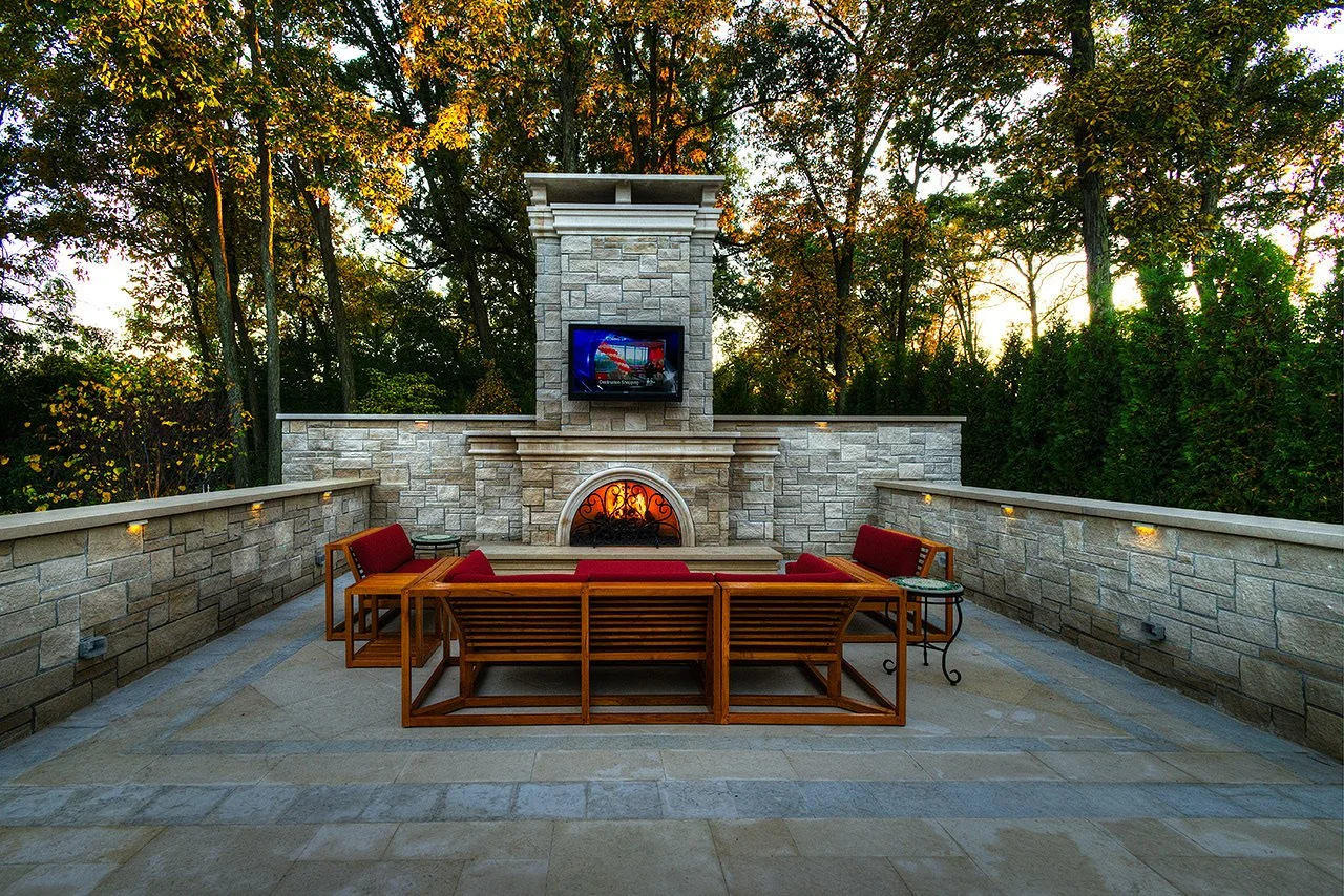 Outdoor patio with stone walls, a built-in fireplace, and a mounted TV under a crown molding at sunset. Contains wooden outdoor seating with red cushions and mixed side tables, surrounded by trees.