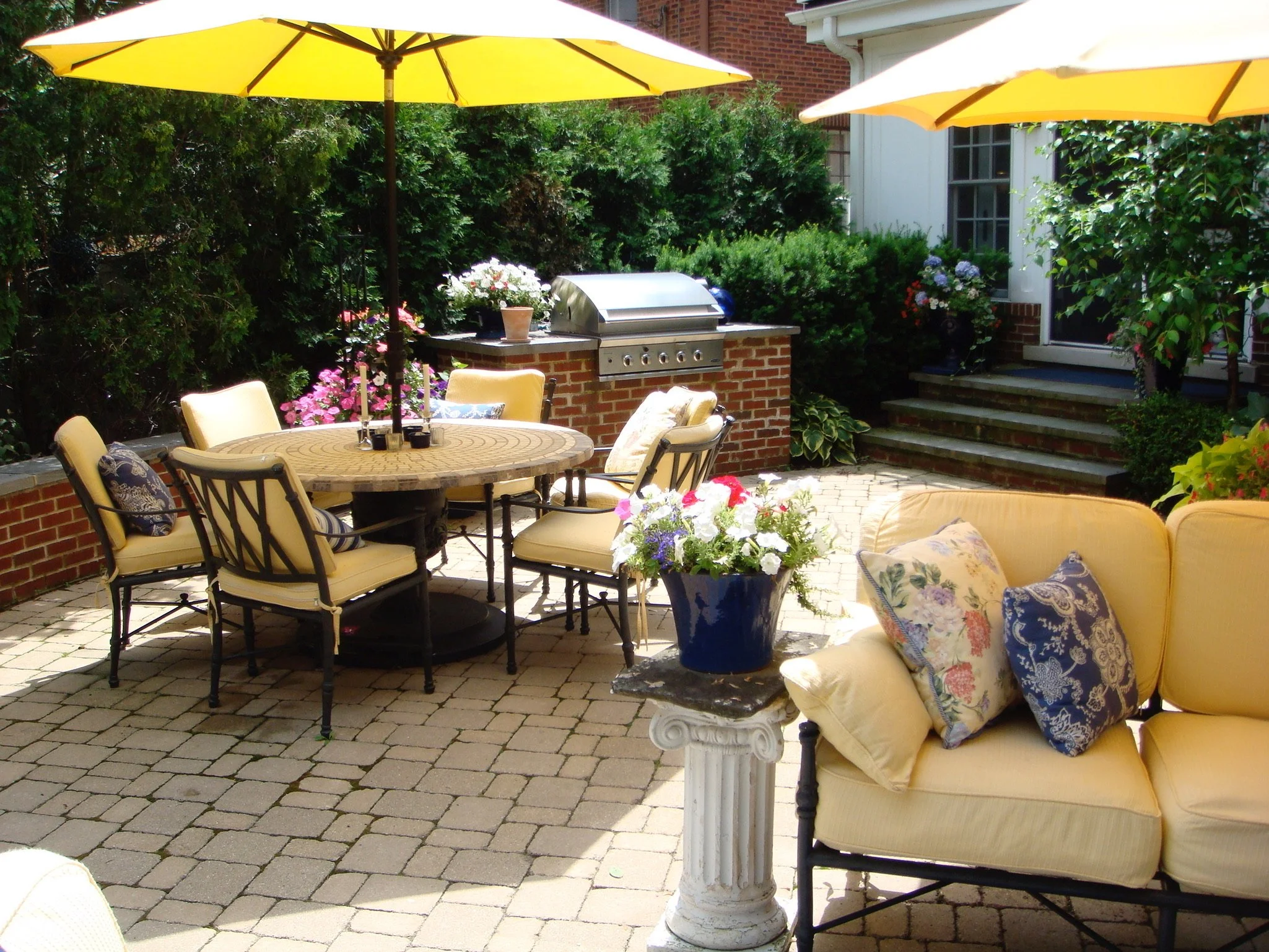 A backyard patio area with a round table and chairs, yellow umbrellas, a built-in grill, potted flowers, and outdoor furniture decorated with patterned pillows, surrounded by greenery and brick walls.