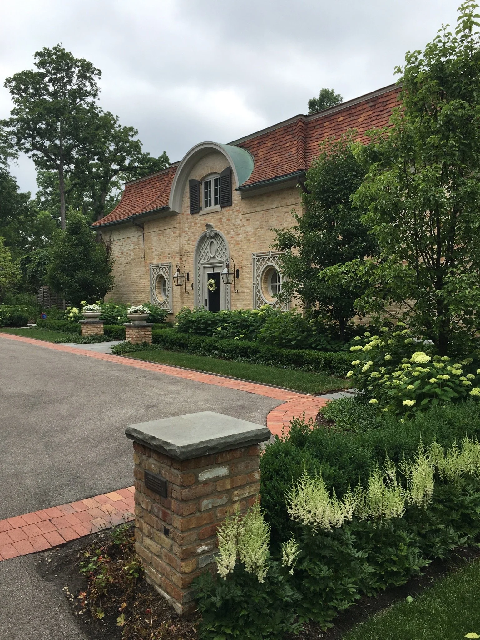 A brick house with a curved roof and black shutters, surrounded by lush greenery, flowers, and a brick border walkway.