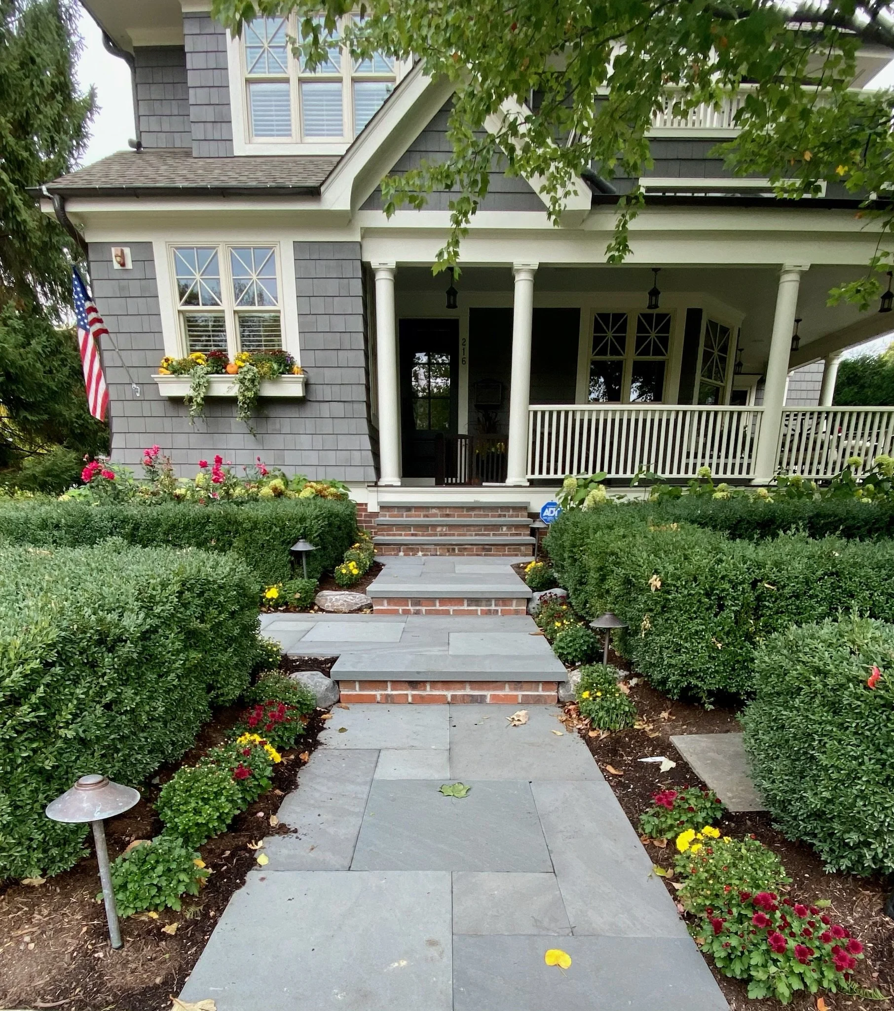 A front porch and walkway leading to a house with a gray and white exterior, surrounded by well-maintained flower beds with yellow and pink flowers, lush green bushes, and trees.