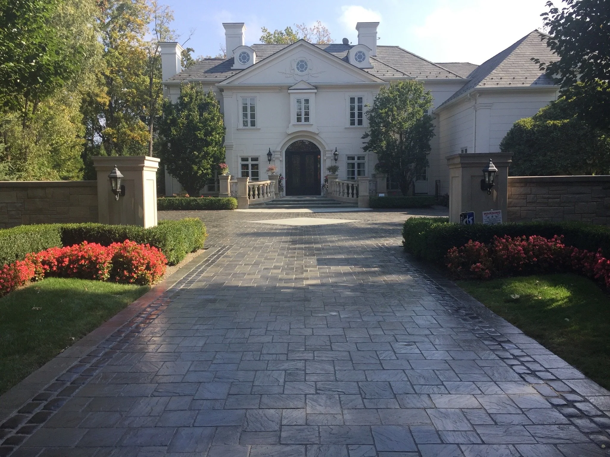 A large white house with a front porch, multiple windows, and a dark door. The house is surrounded by trees and a stone-paved driveway with neatly trimmed bushes and flower beds.