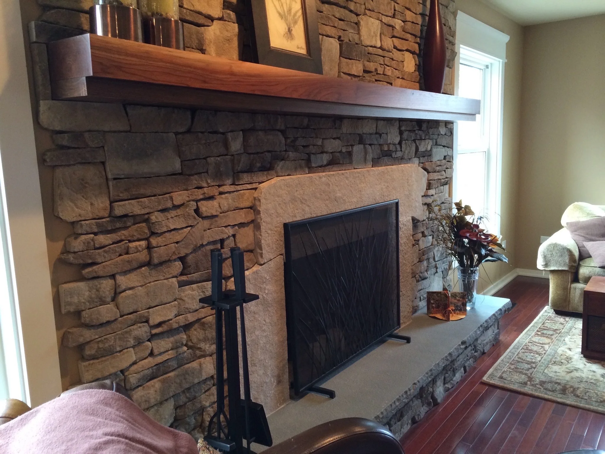 Living room with a stone fireplace, decorated with a framed picture, tall vases, and potted plants on a mantel. There is a window to the right, curtains, a sofa with cushions, and a rug on wooden floor.