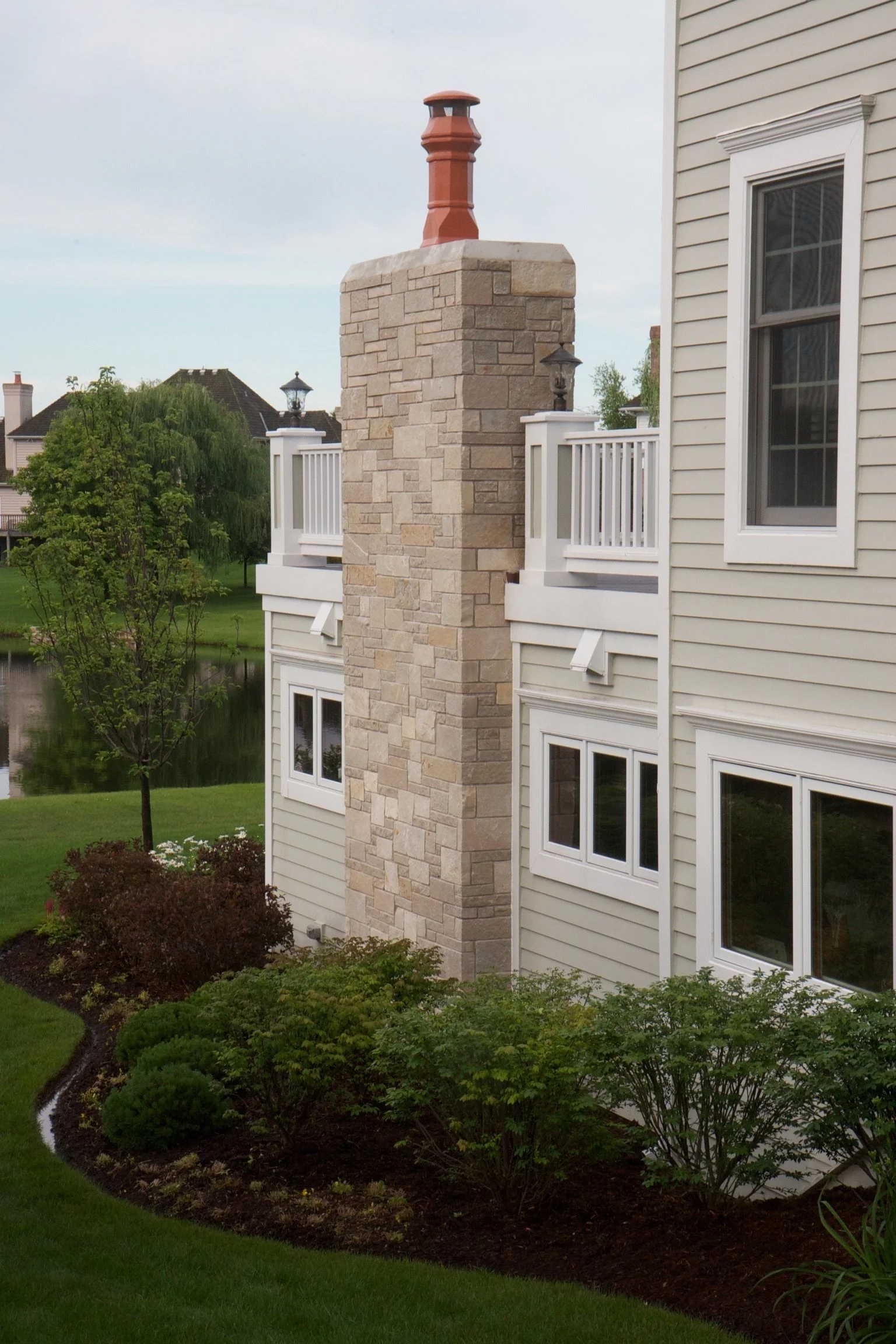 A house exterior with a stone chimney, beige siding, white-framed window, a white balcony railing, and landscaped garden with bushes and grass.