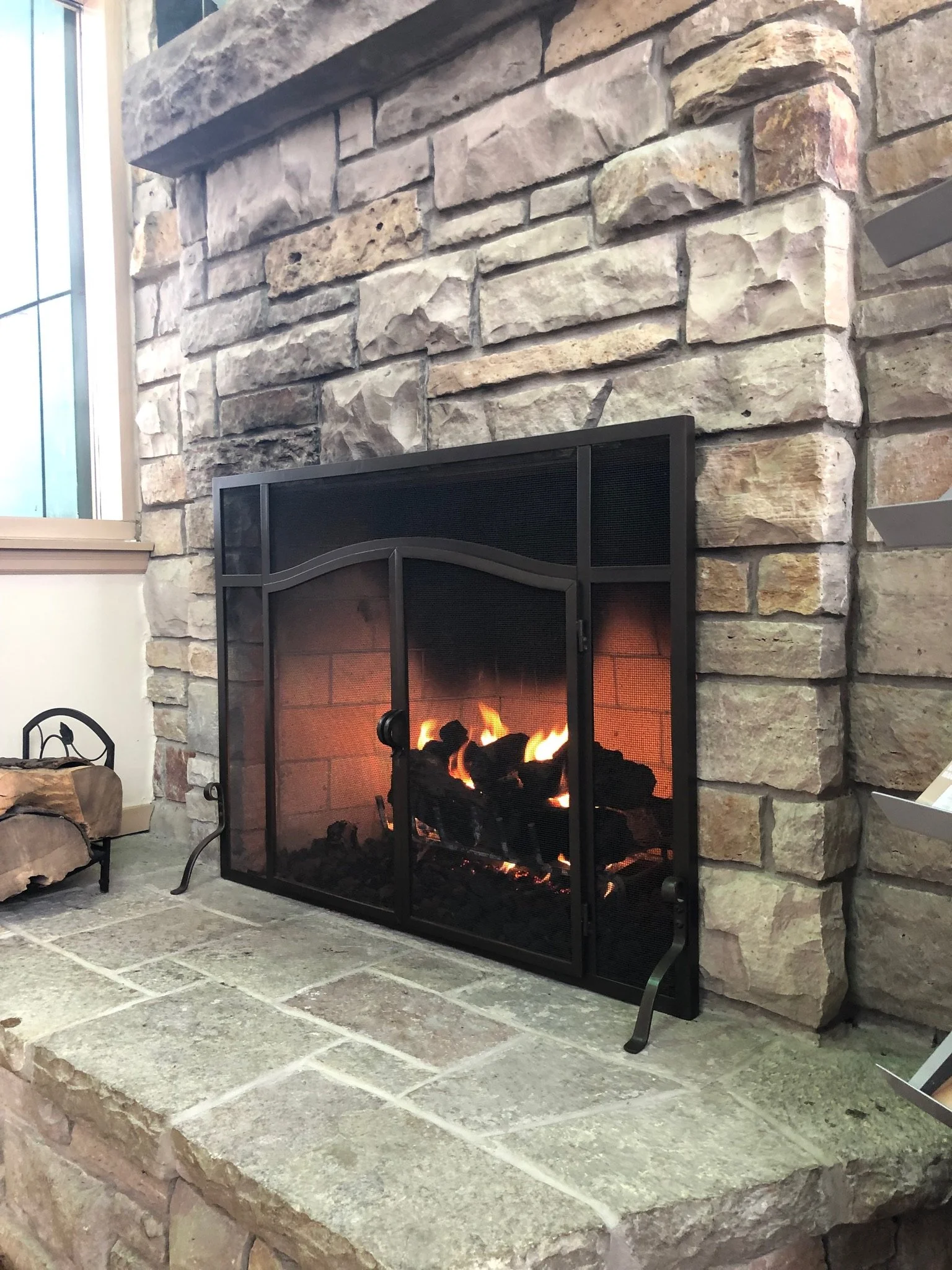 A fireplace with a stone surround, showing glowing logs inside a black metal frame with a glass front, located on a stone hearth.