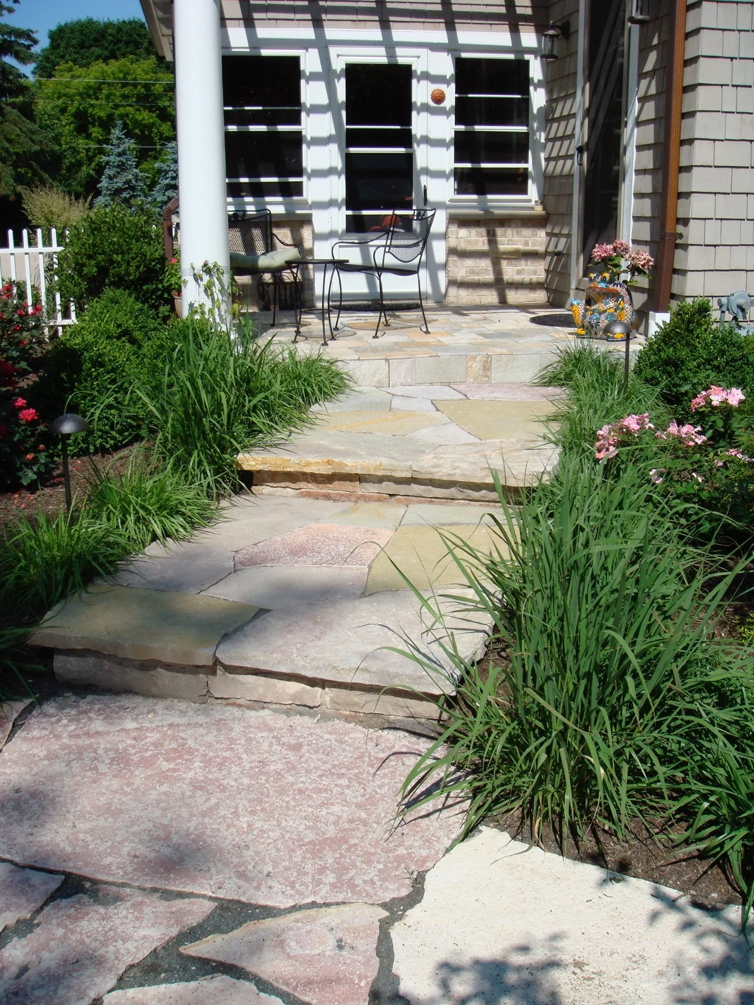 A stone porch with steps leads to a house with beige siding, white trim, and three windows. There is an outdoor seating area with a small black iron table and chairs, a decorative frog garden statue with pink flowers, and various green plants and bus