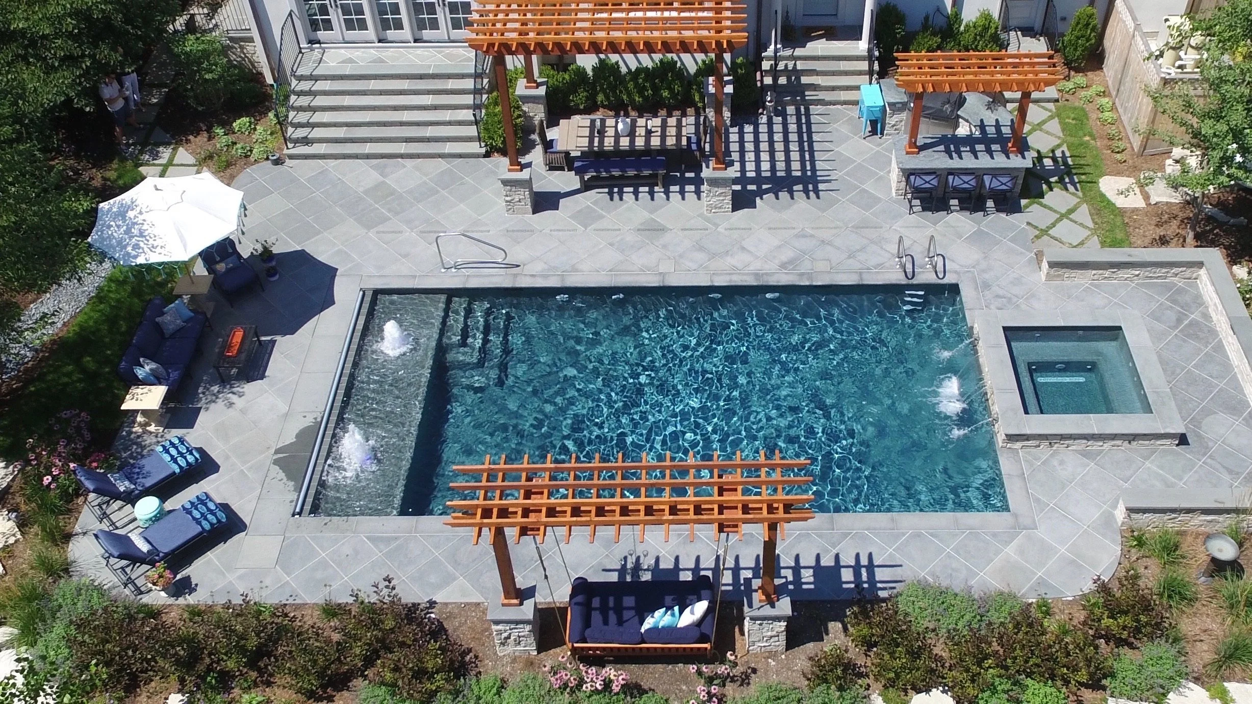 Aerial view of a backyard with a swimming pool, lounge chairs, umbrellas, a hot tub, and wooden pergolas surrounded by greenery and plants.