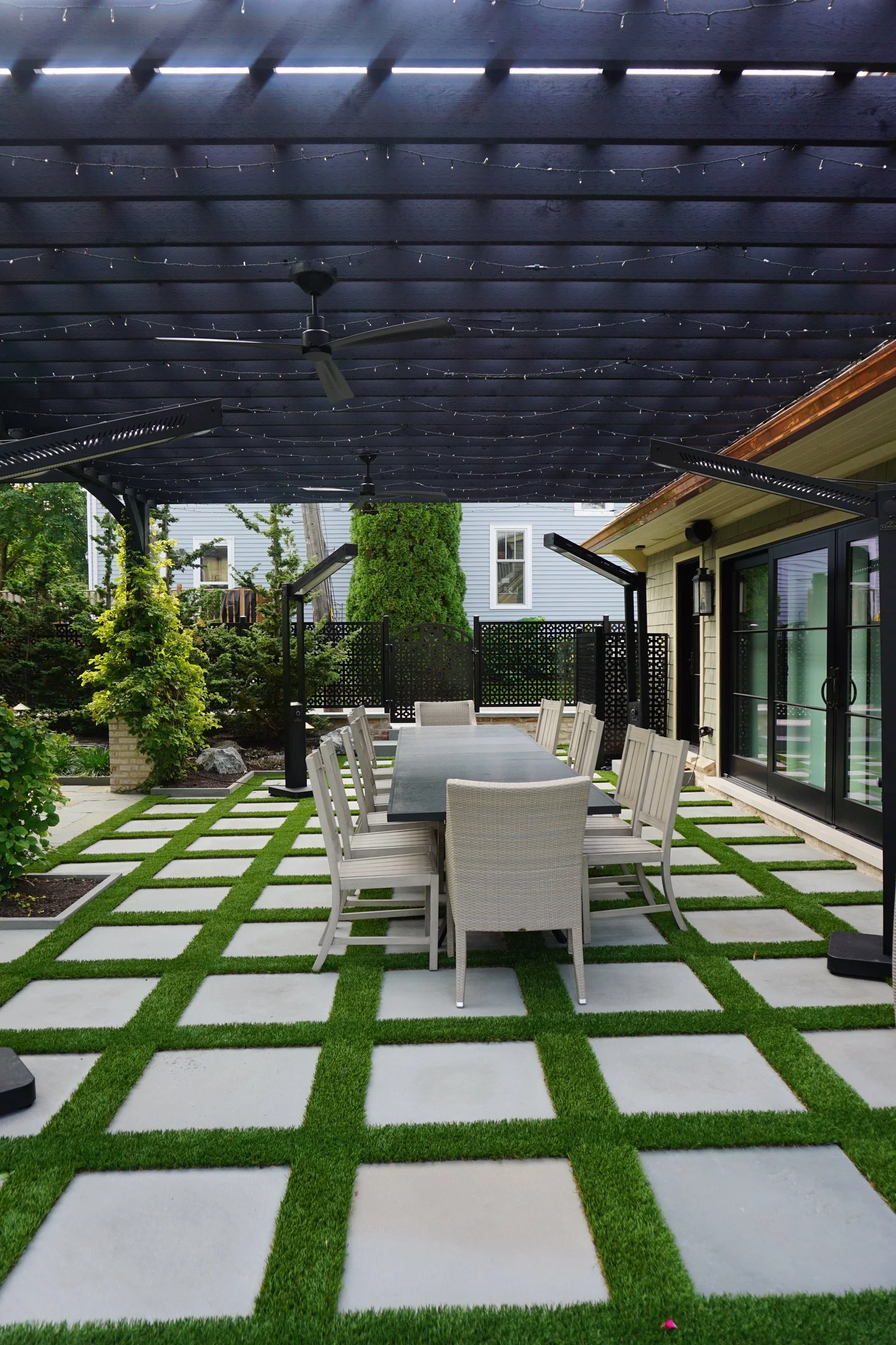 Outdoor patio area with a black and white checkered tile and artificial grass flooring, large dining table with white chairs, a black pergola with string lights, ceiling fans, and neighboring houses in the background.