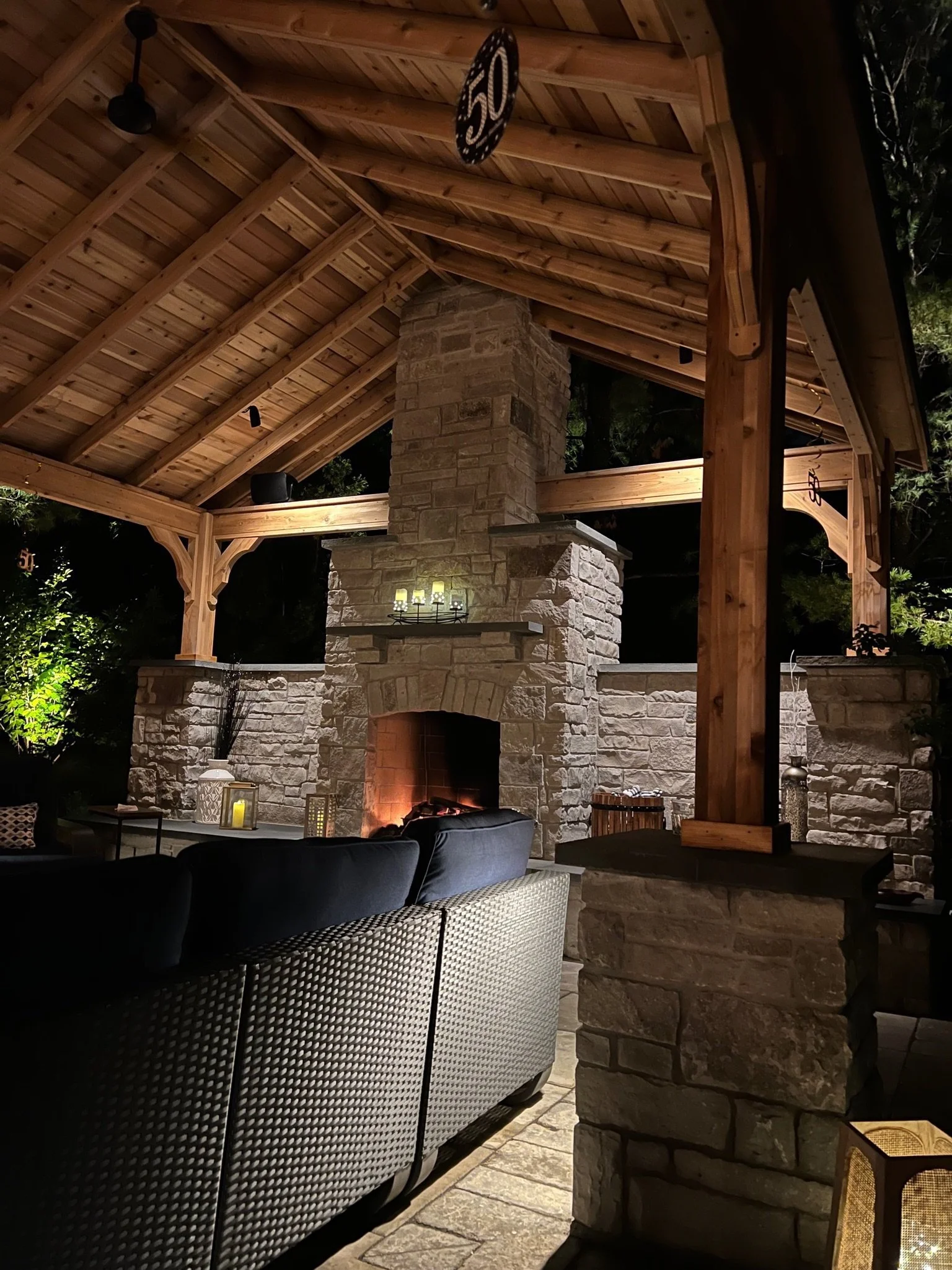 Nighttime outdoor patio with a stone fireplace, wooden roof Beams, and modern chairs.