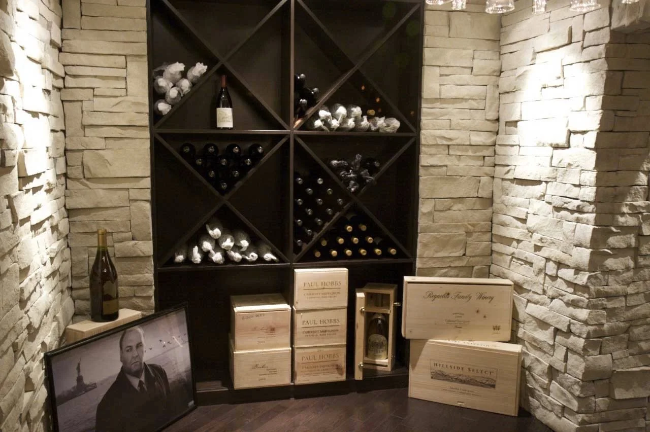 Wine bottles stored on a black wine rack against a white brick wall. Some wine boxes and a framed photo are placed on the floor in front of the rack, with additional boxes and a small lantern on the right.