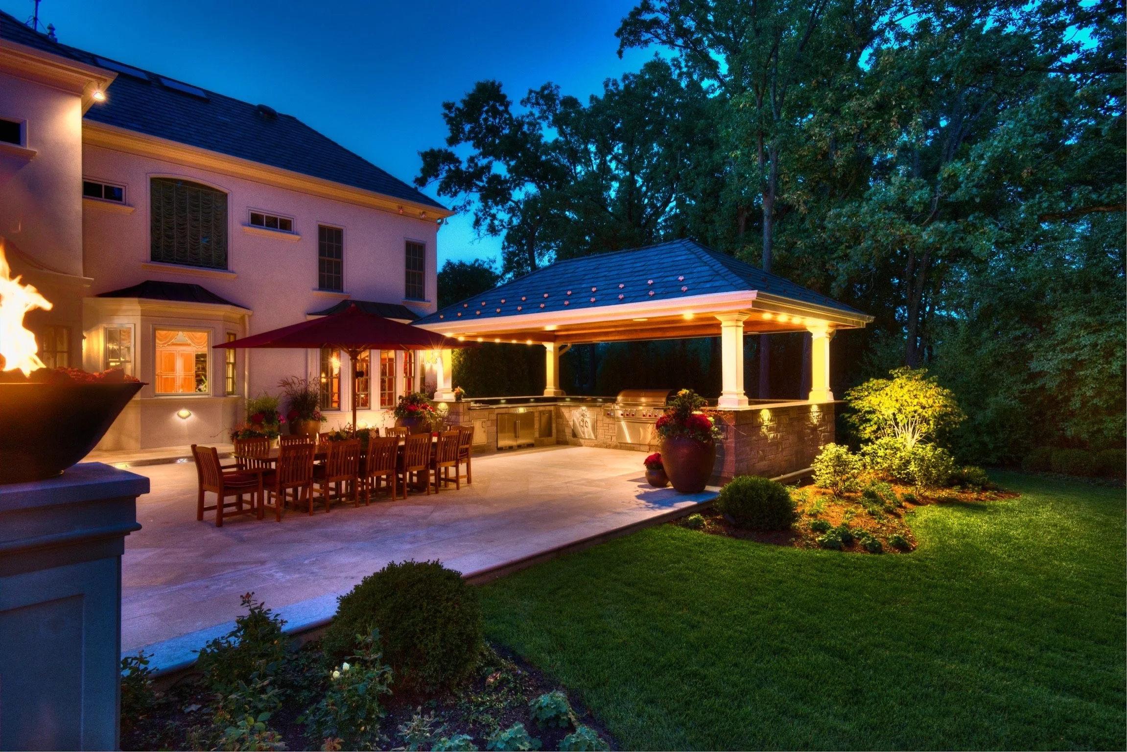 Backyard patio at dusk with outdoor dining area, fireplace, and landscaped garden surrounded by tall trees.