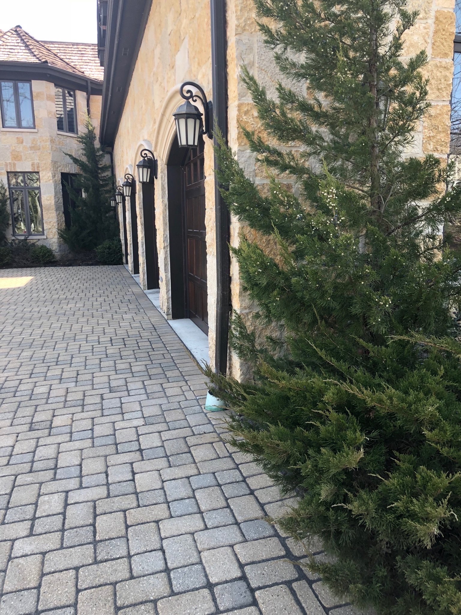 Exterior view of a beige stone building with black outdoor wall lanterns, a paved walkway, and a green pine tree in the foreground.