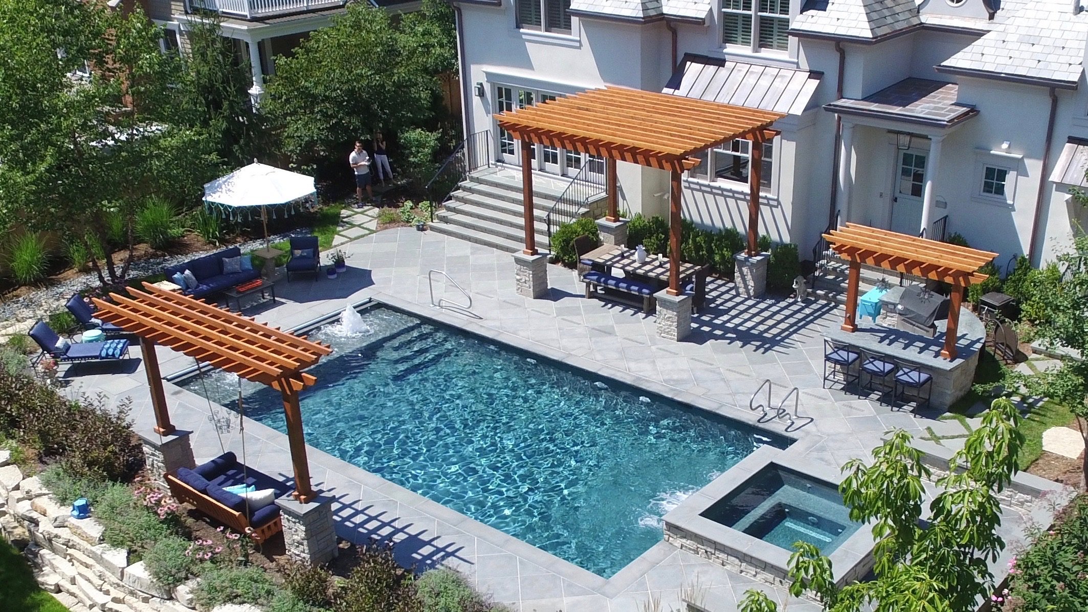 A backyard with a swimming pool, shaded pergolas, outdoor furniture, and surrounding greenery.