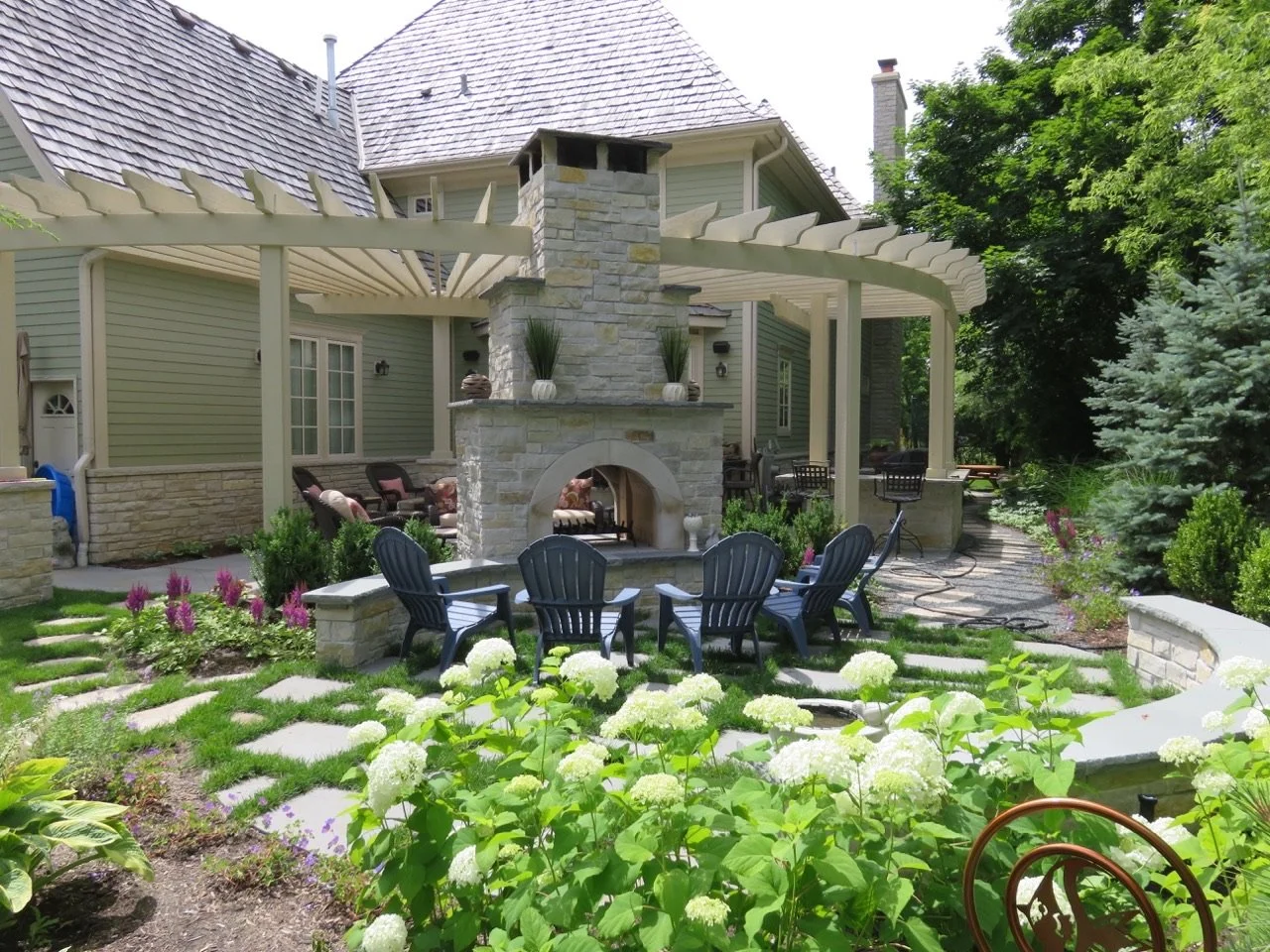 Backyard patio with stone fireplace, Adirondack chairs, garden with white hydrangeas, stone pathway, and a pergola attached to a house with green siding and a shingled roof.