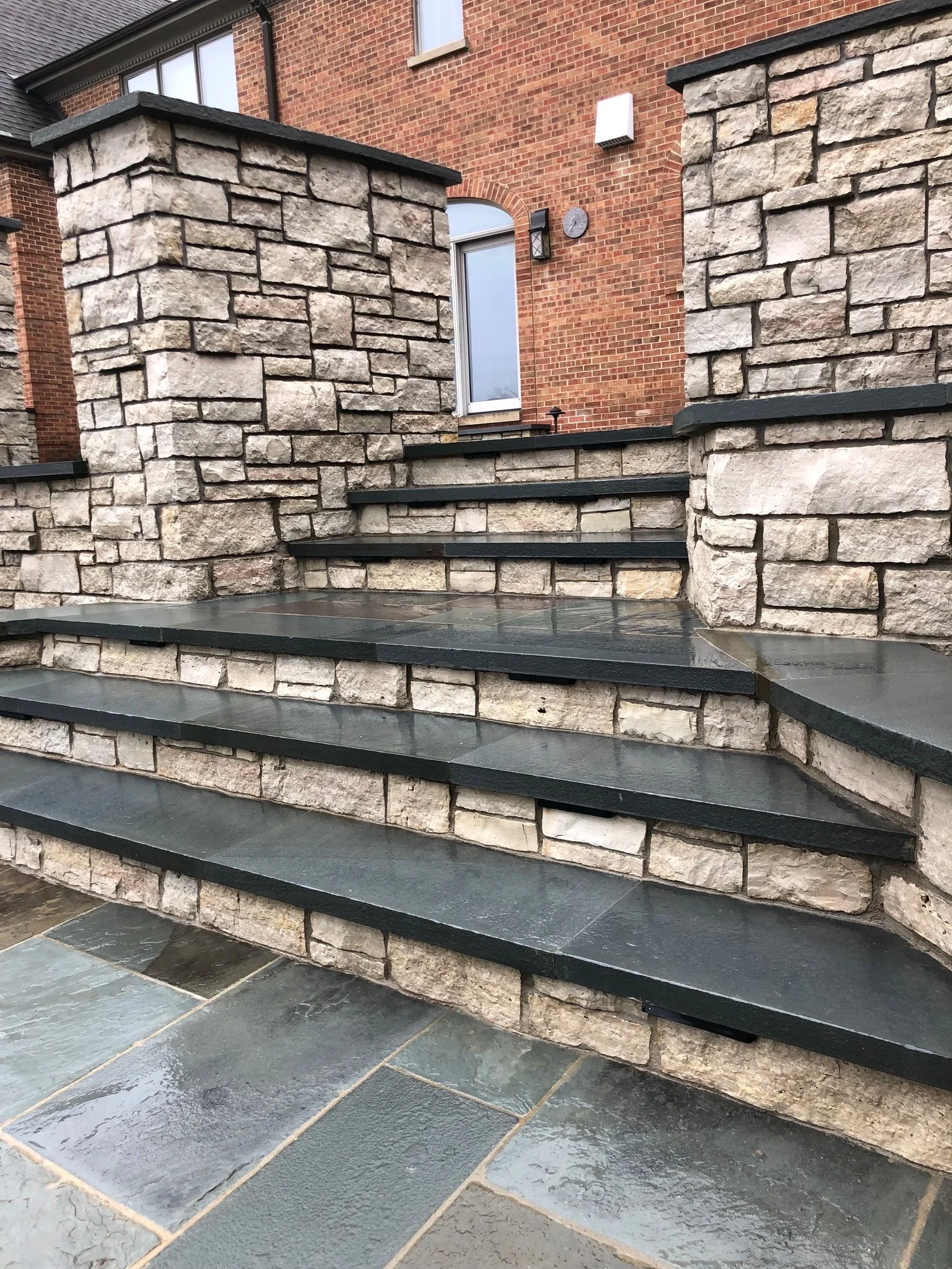 Stone steps with dark gray tiles leading up to a brick house with a large window.