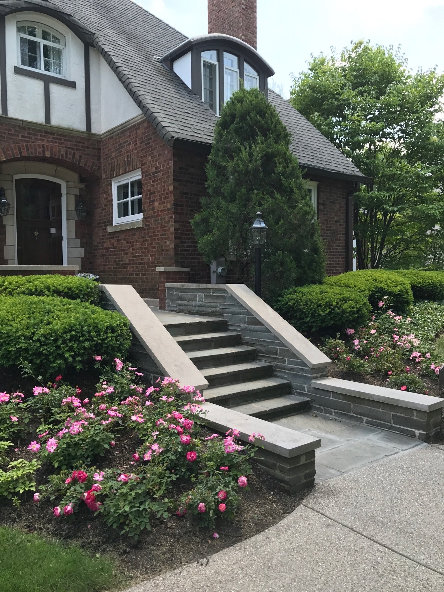 Front yard of a brick house with steps leading to the door, surrounded by pink and red flowers, green bushes, and a large tree, with a lamp post near the steps.