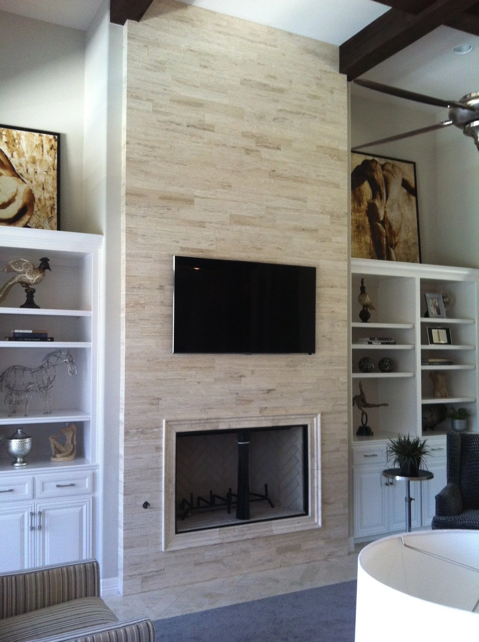 Living room with beige stone fireplace, black TV mounted above fireplace, white shelving units on both sides with decorative items, dark wood beams on ceiling, artwork on walls, gray rug, and a round white table with a plant in a black pot.