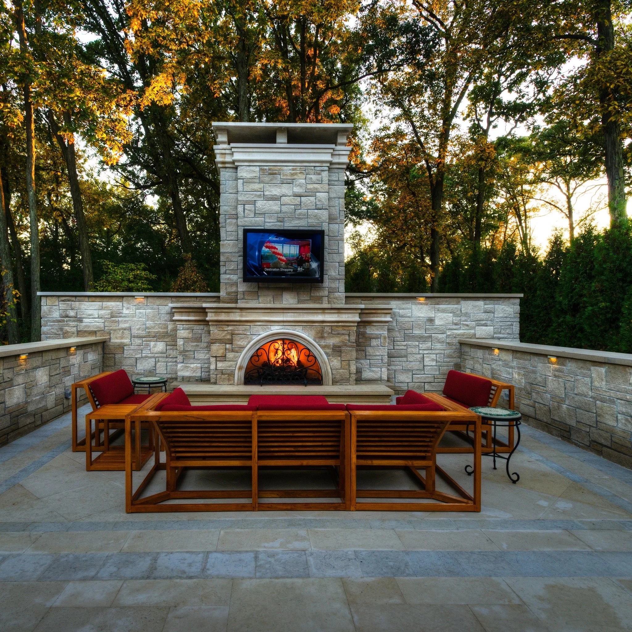 An outdoor scene with a stone fireplace, a mounted television, and wooden seating, surrounded by trees with autumn leaves.