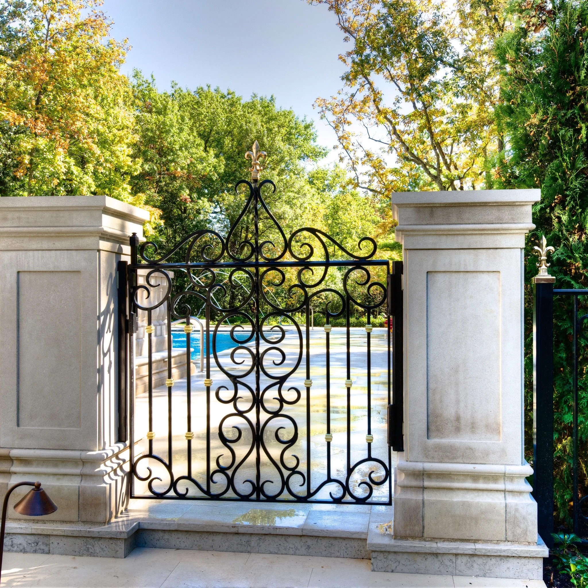 A black ornate iron gate with gold accents, flanked by two stone pillars, leading to a backyard pool area with trees in the background, and the sky visible overhead.